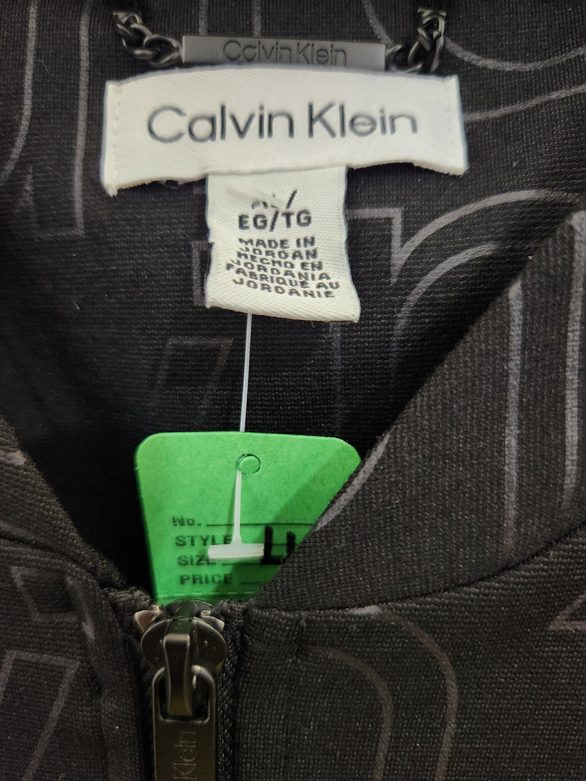 Close-up of a Calvin Klein zippered garment showing the inner neck label, a green size tag, and part of the dark patterned fabric.