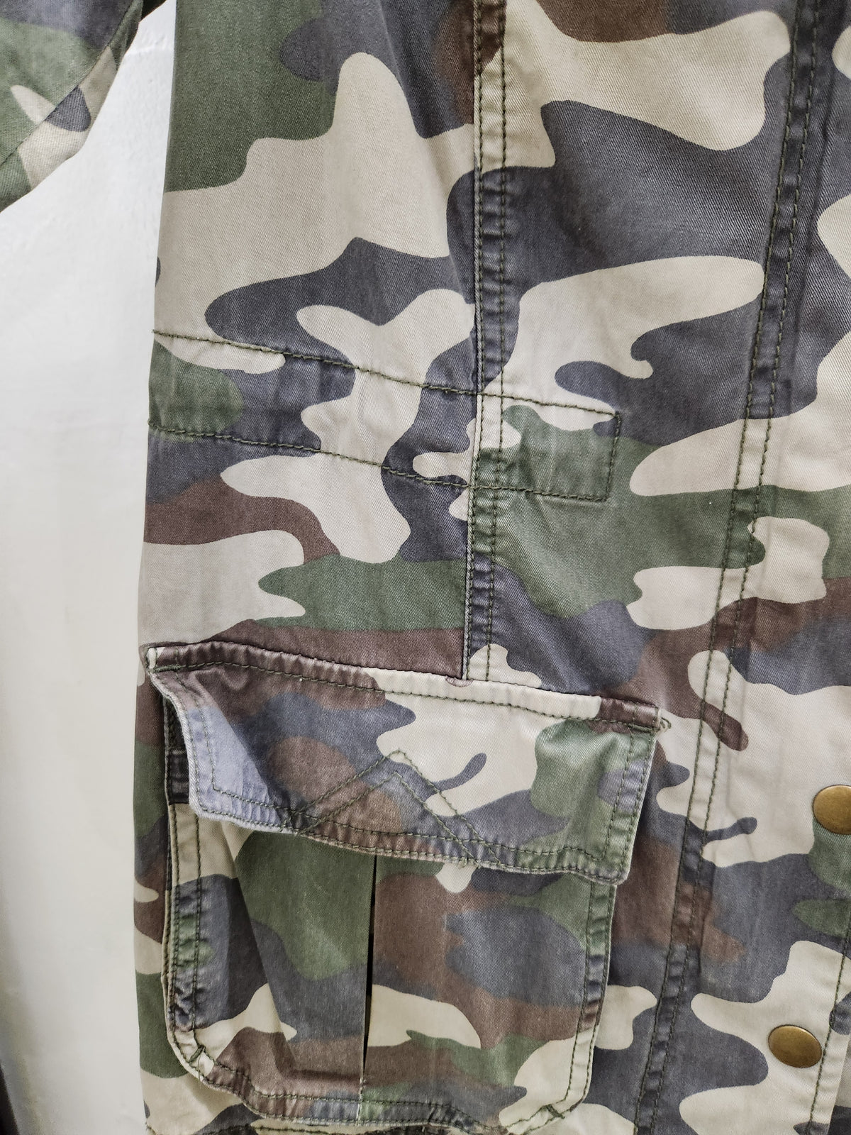 Close-up of a camouflage jacket with a front flap pocket and metal snap buttons, shown against a white background.