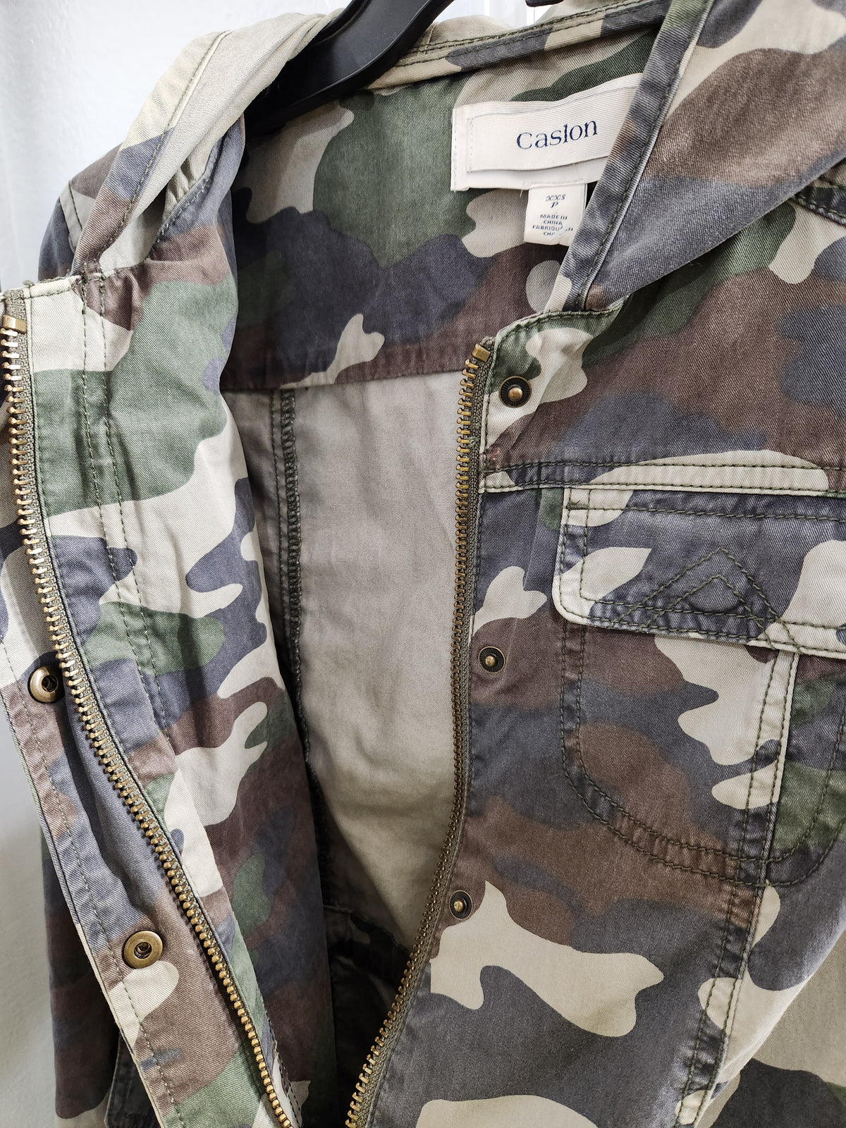 A close-up of a camouflage jacket hanging on a black hanger, showing the interior lining, brass zipper, and a visible Casion brand label.