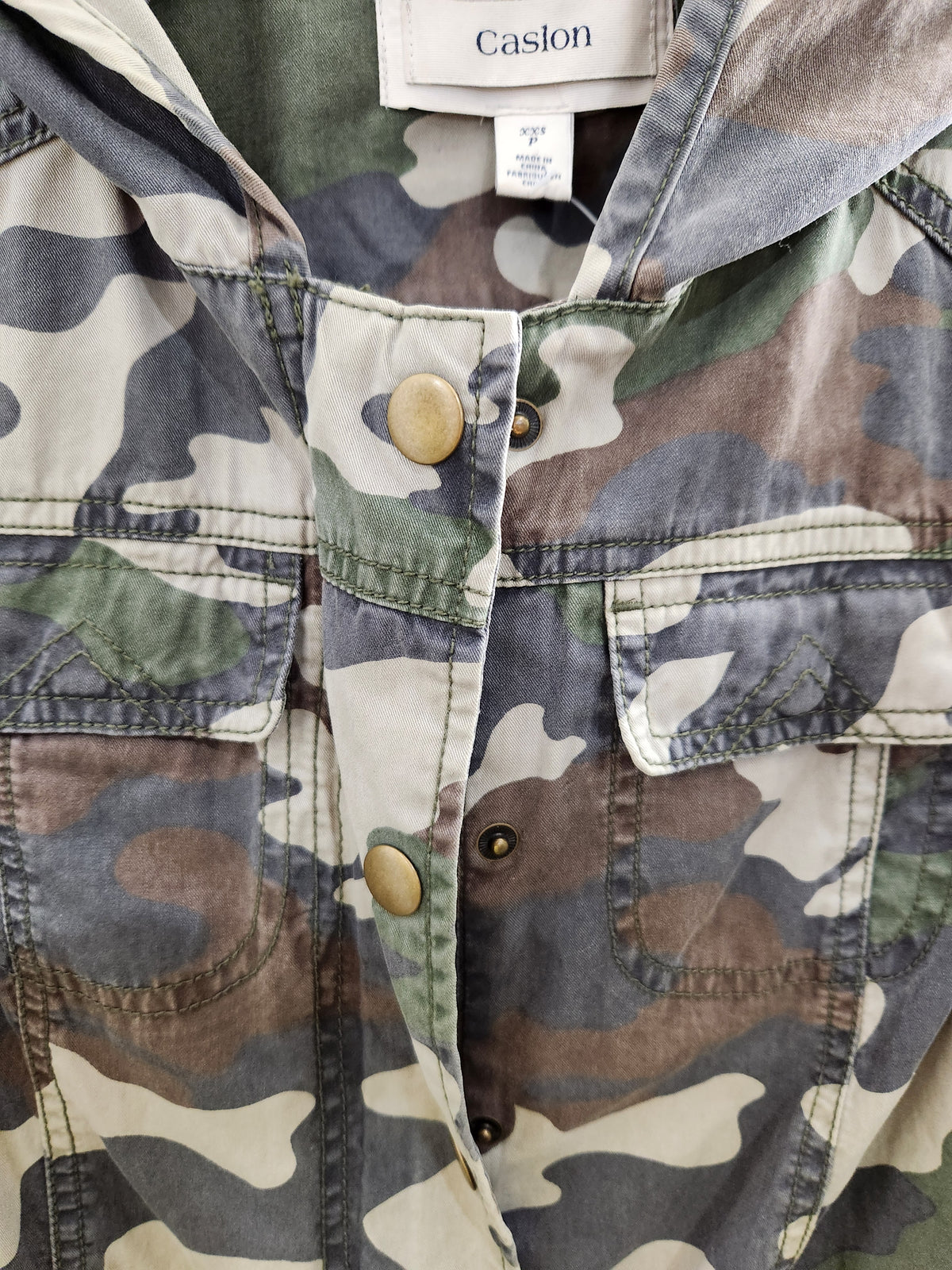Close-up of a camouflage jacket with a hood, featuring a muted green, brown, beige, and gray camo pattern, brass buttons, and a visible Caslon brand label.