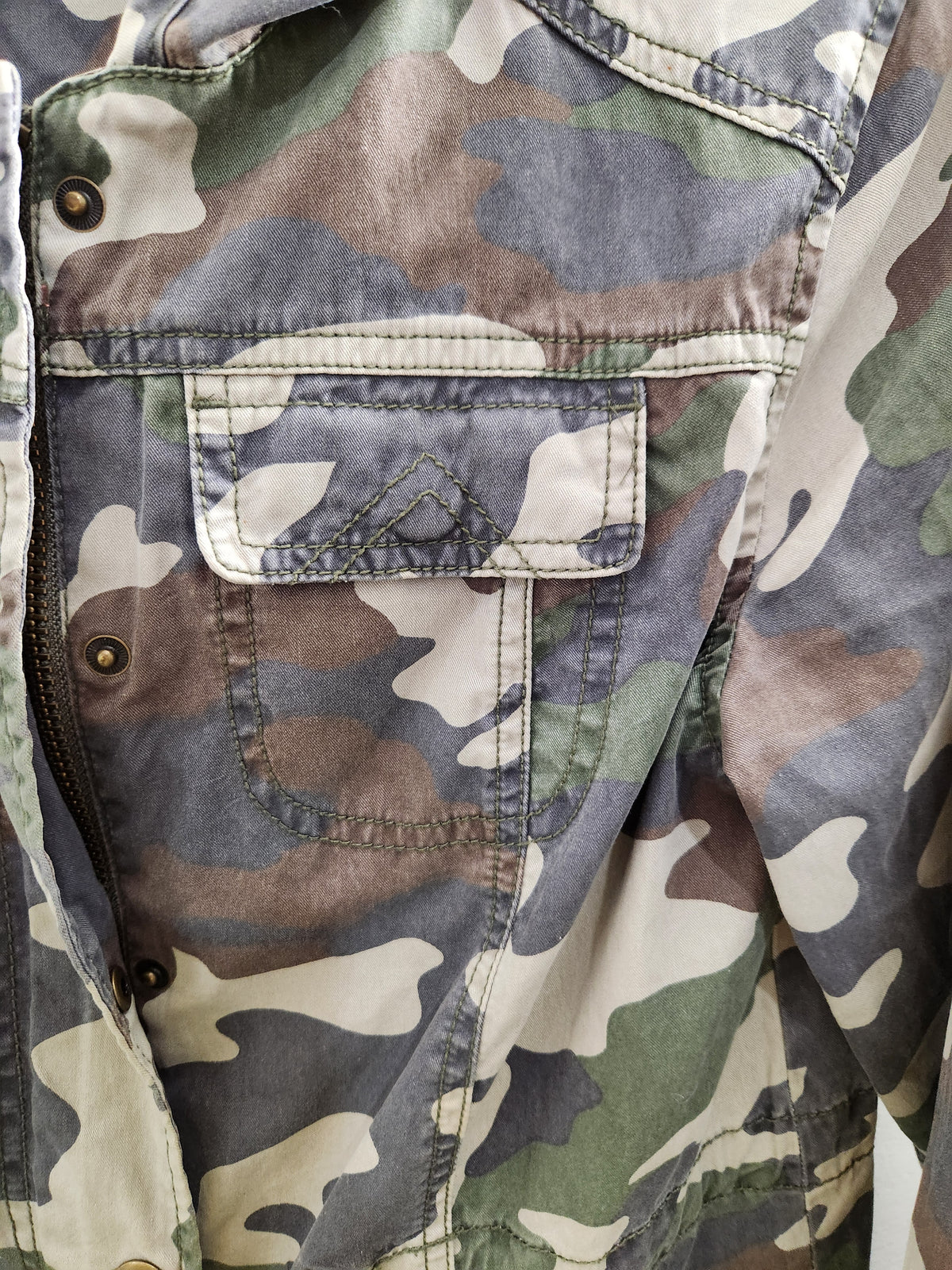 Close-up of a camouflage jacket with a flap chest pocket, showing muted green, gray, beige, and brown tones.