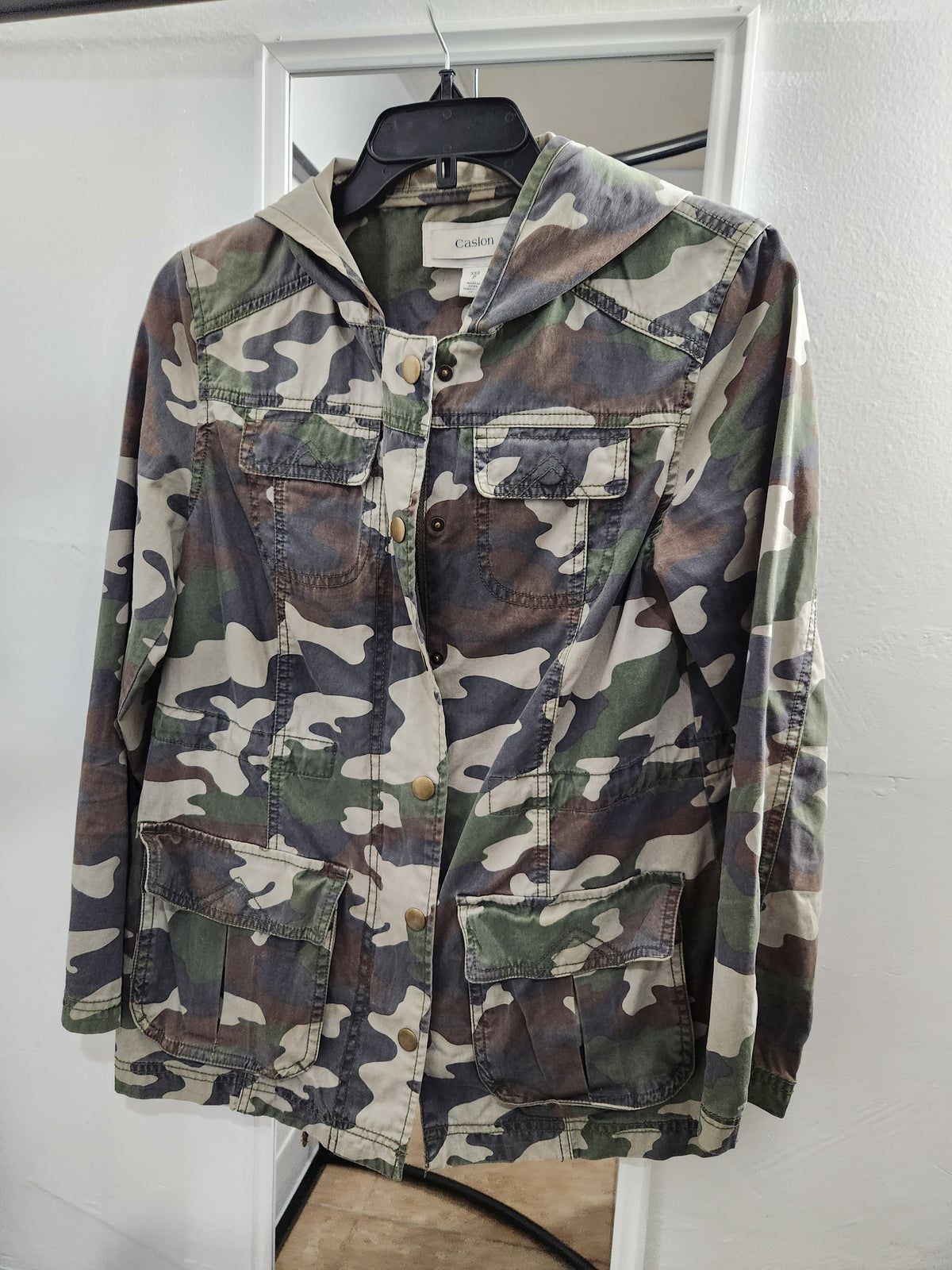 A camouflage utility jacket hanging on a black hanger against a white wall and mirror background. The jacket features a classic woodland camo pattern with green, brown, beige, and black tones, multiple front pockets, and a button closure.