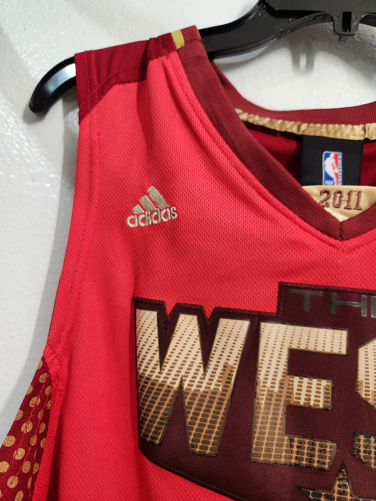 A close-up of a red Adidas basketball jersey hanging on a black hanger against a white wall. The jersey features gold and dark maroon lettering, a gold Adidas logo, and an NBA 2011 patch on the inside neck label.