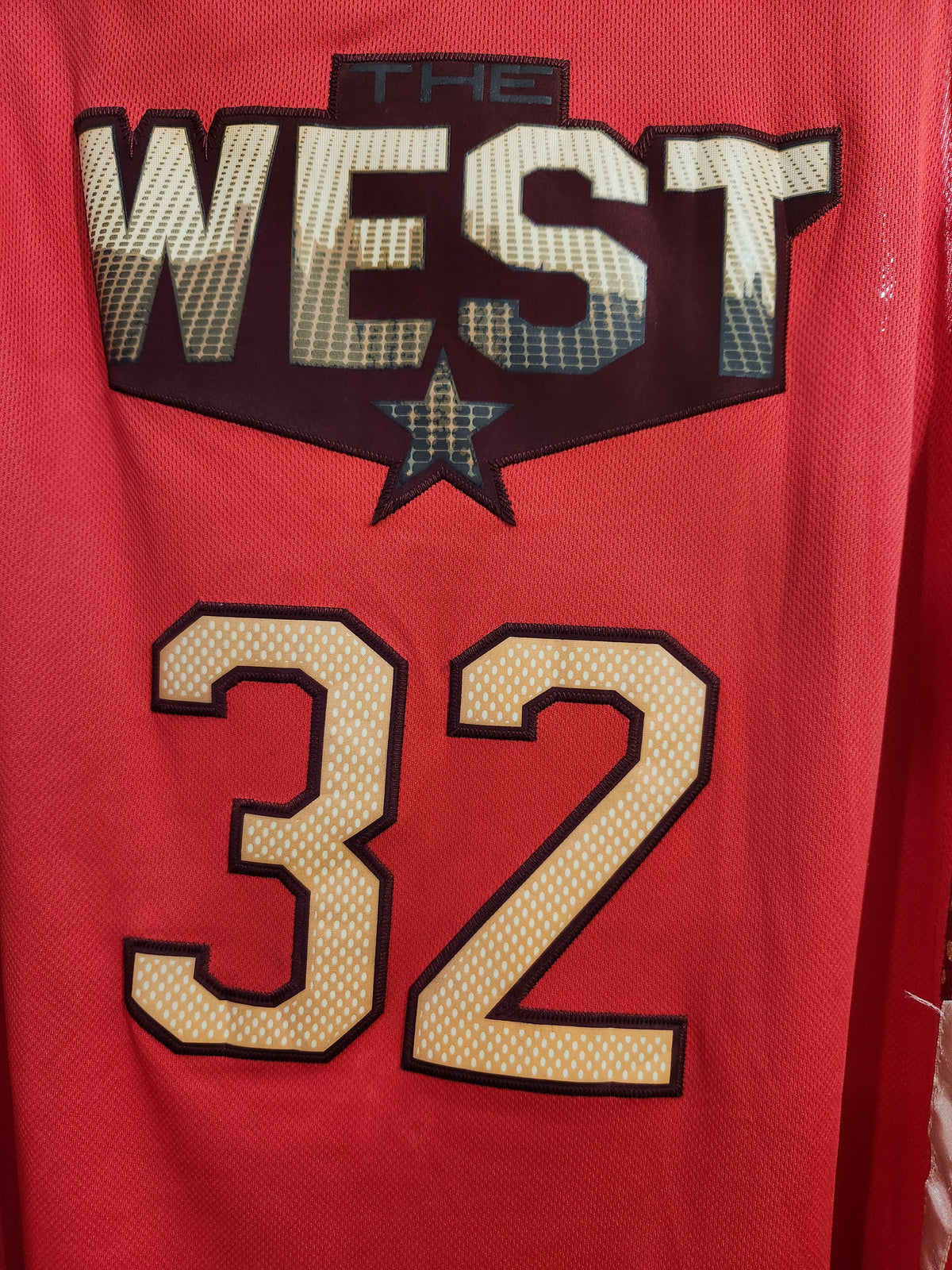 Close-up of the back of a red mesh basketball jersey featuring 'THE WEST' in large patterned letters above a star and the number '32' in beige with black outlines.