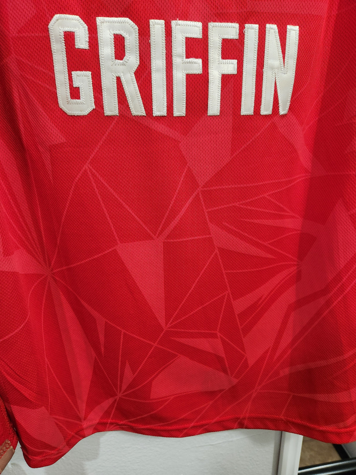 Close-up of a red athletic jersey with the name GRIFFIN in large white block letters across the upper back, over a subtle geometric pattern.