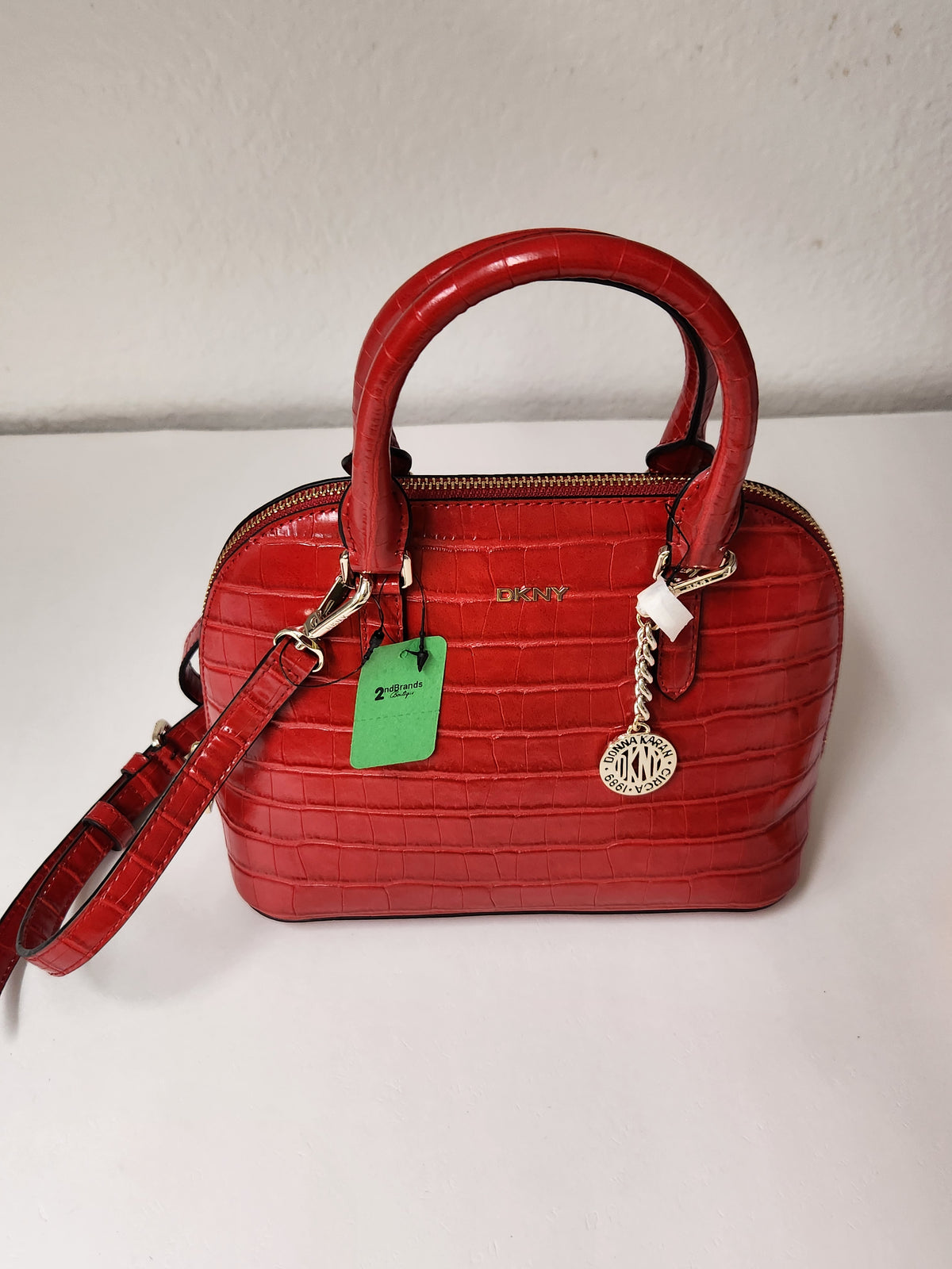 A red crocodile-embossed leather handbag with double top handles, a detachable shoulder strap, silver-tone hardware, and a green product tag, photographed on a white surface against a white wall.
