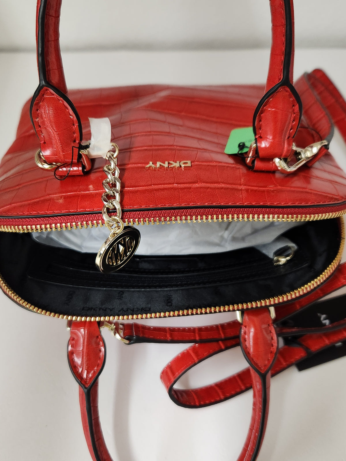 A close-up of a red crocodile-embossed leather handbag by DKNY, shown open to reveal a black interior. The bag features gold-tone hardware, a hanging circular DKNY charm, and a small green hangtag.