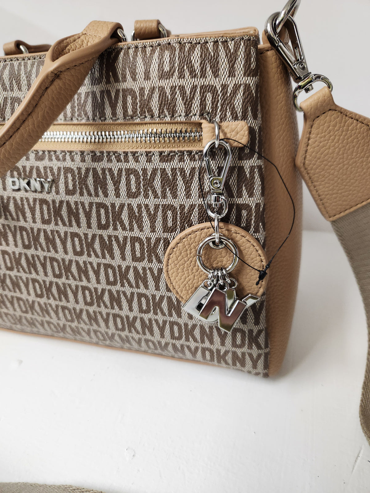 A close-up of a DKNY crossbody bag in beige and brown monogram canvas with tan leather trim, a silver zipper, and a hanging keychain charm with a silver 'N' initial.