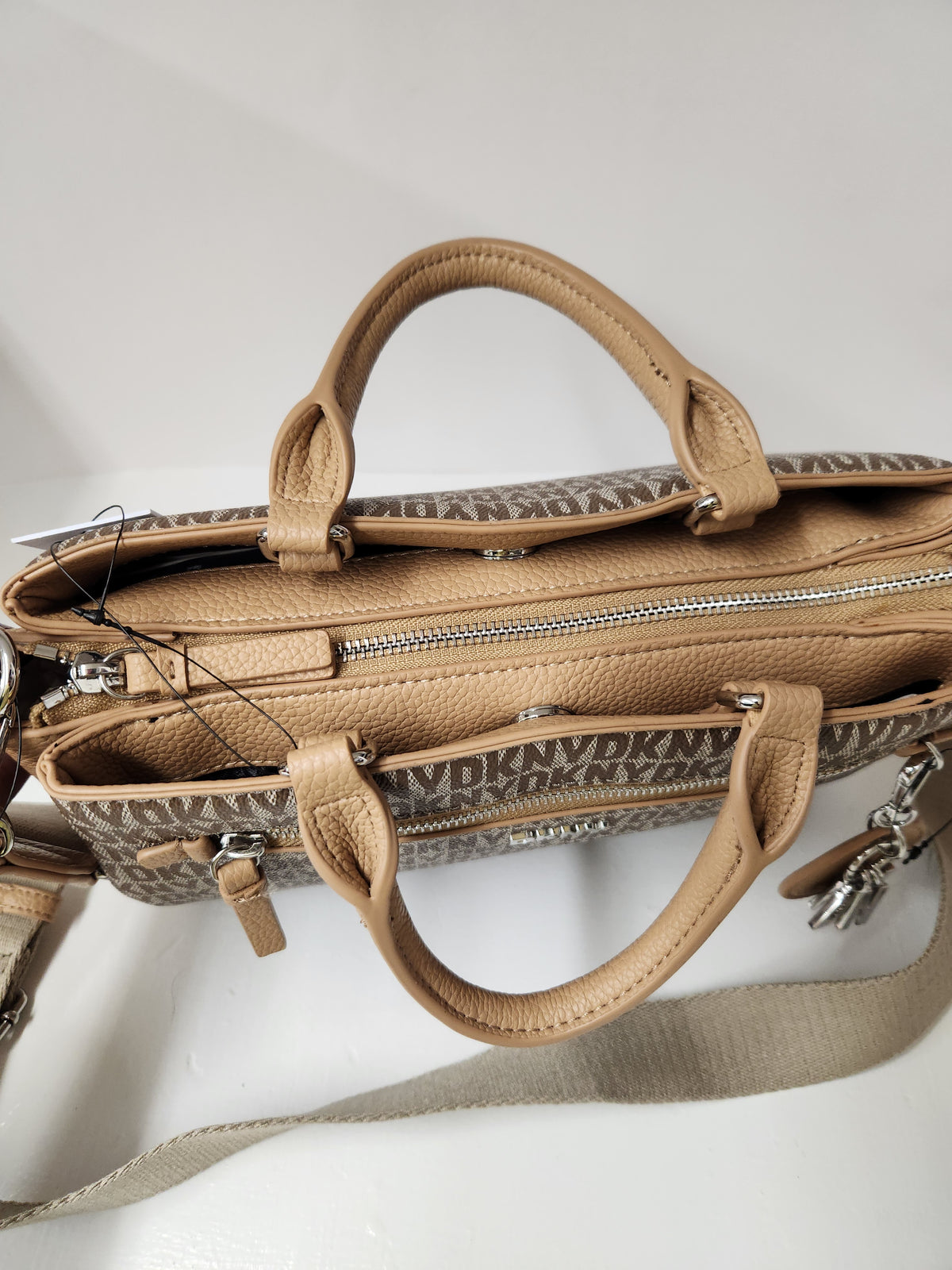 A beige and brown monogram handbag with tan leather handles and a matching shoulder strap, shown on a light gray background.