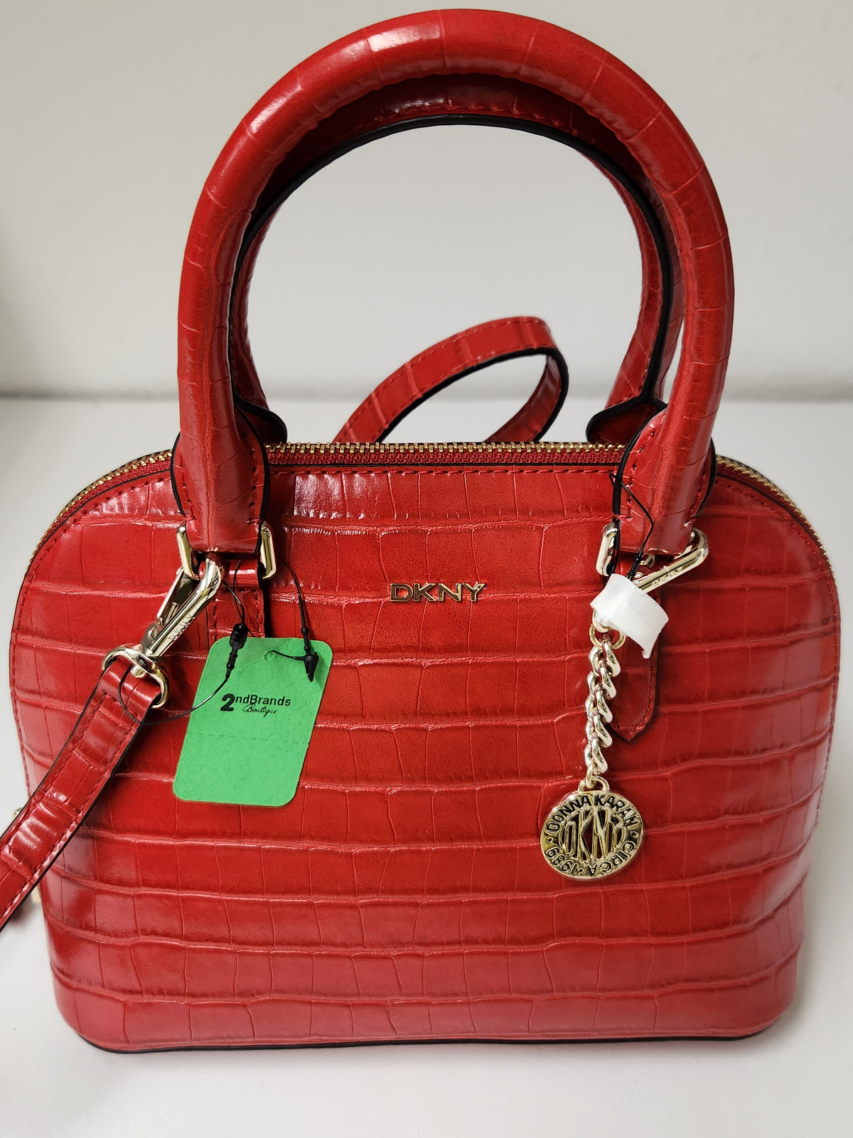 A red crocodile-embossed leather handbag with top handles, a gold-tone DKNY logo on the front, a green product tag, and a hanging metal charm, photographed against a plain light background.