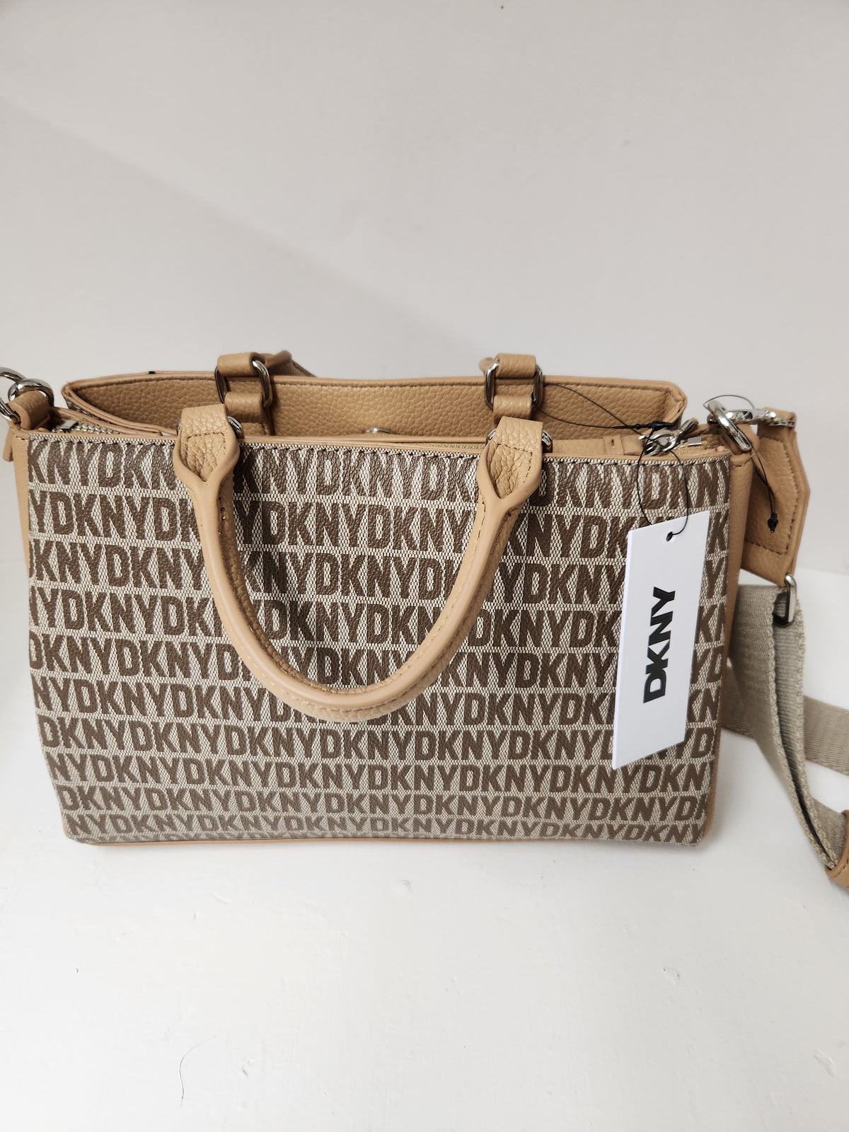 A DKNY tote bag in beige and brown with an allover monogram pattern, featuring tan leather handles and a white hanging DKNY tag.