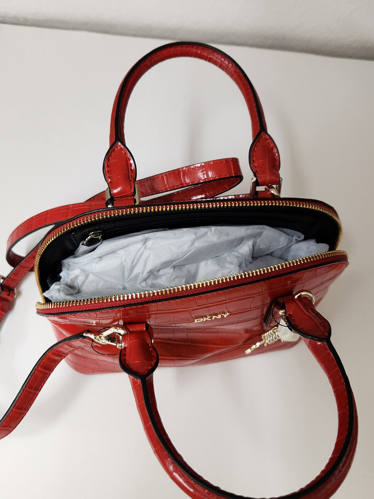 A red crocodile-embossed leather handbag shown open, revealing a light gray protective plastic insert inside. The bag features double top handles, a gold-tone zipper, and a small gold DKNY logo on the front.