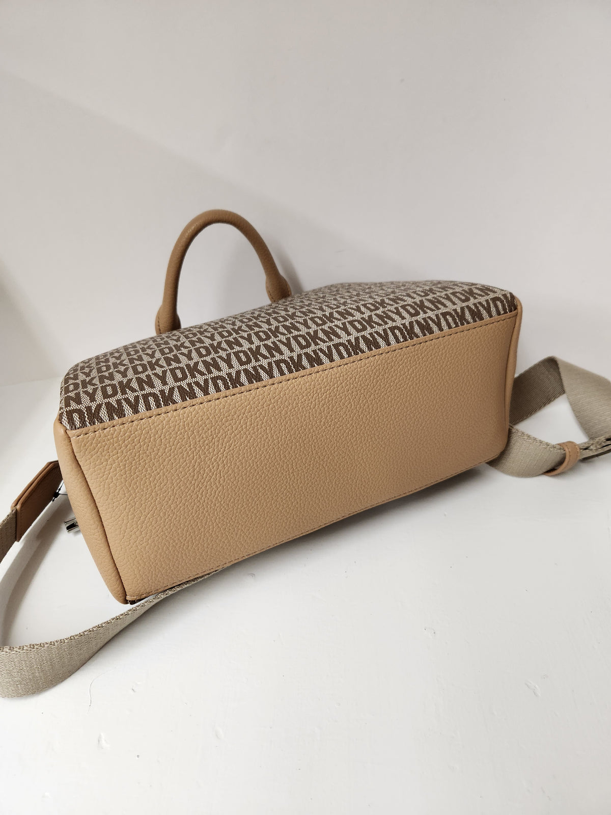 A tan pebbled leather handbag with a brown monogram patterned top panel and a matching beige shoulder strap, photographed on a plain light background.