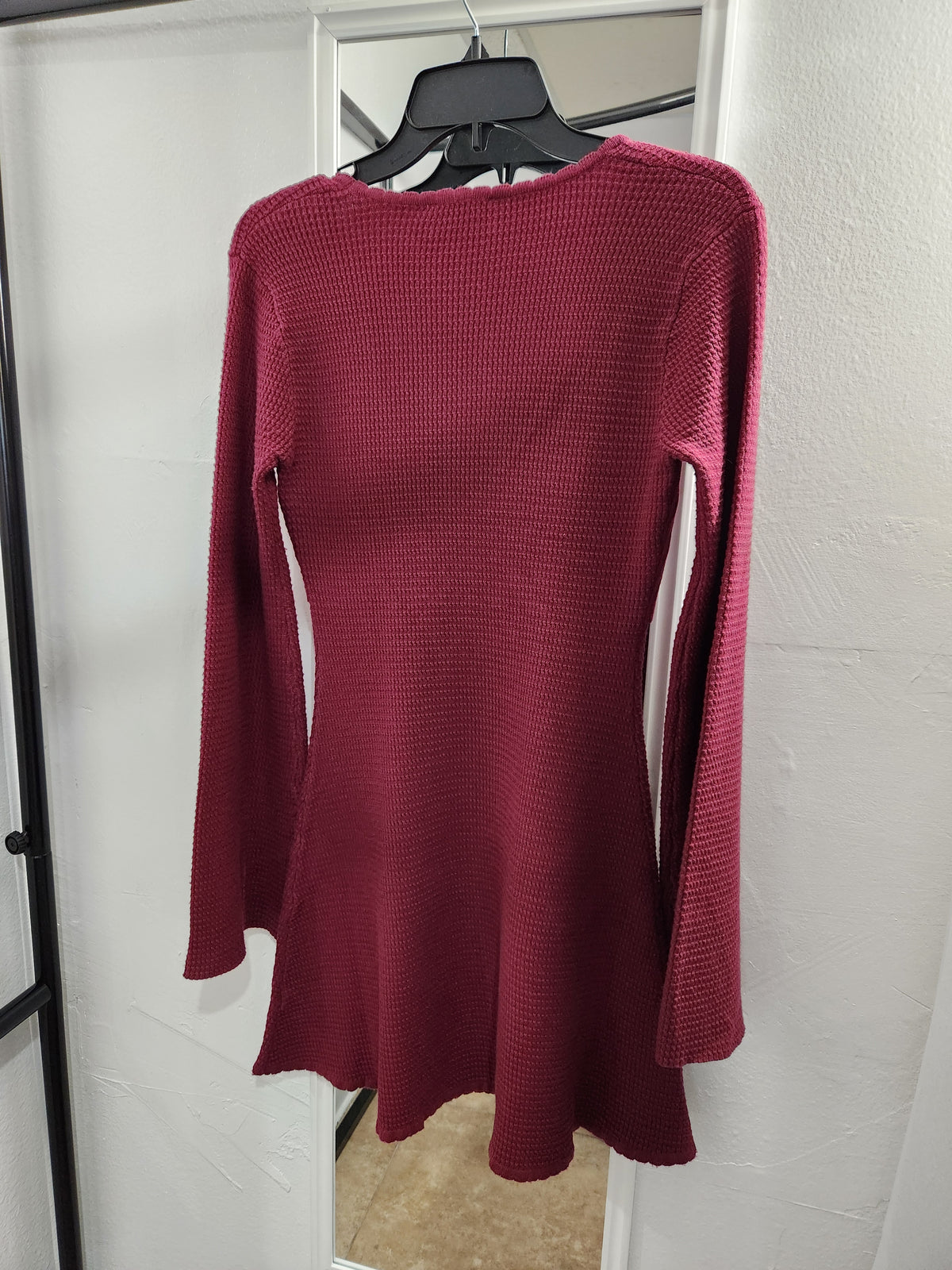 A long-sleeve ribbed knit mini dress in deep burgundy red, hanging on a black hanger in front of a white wall and mirror.