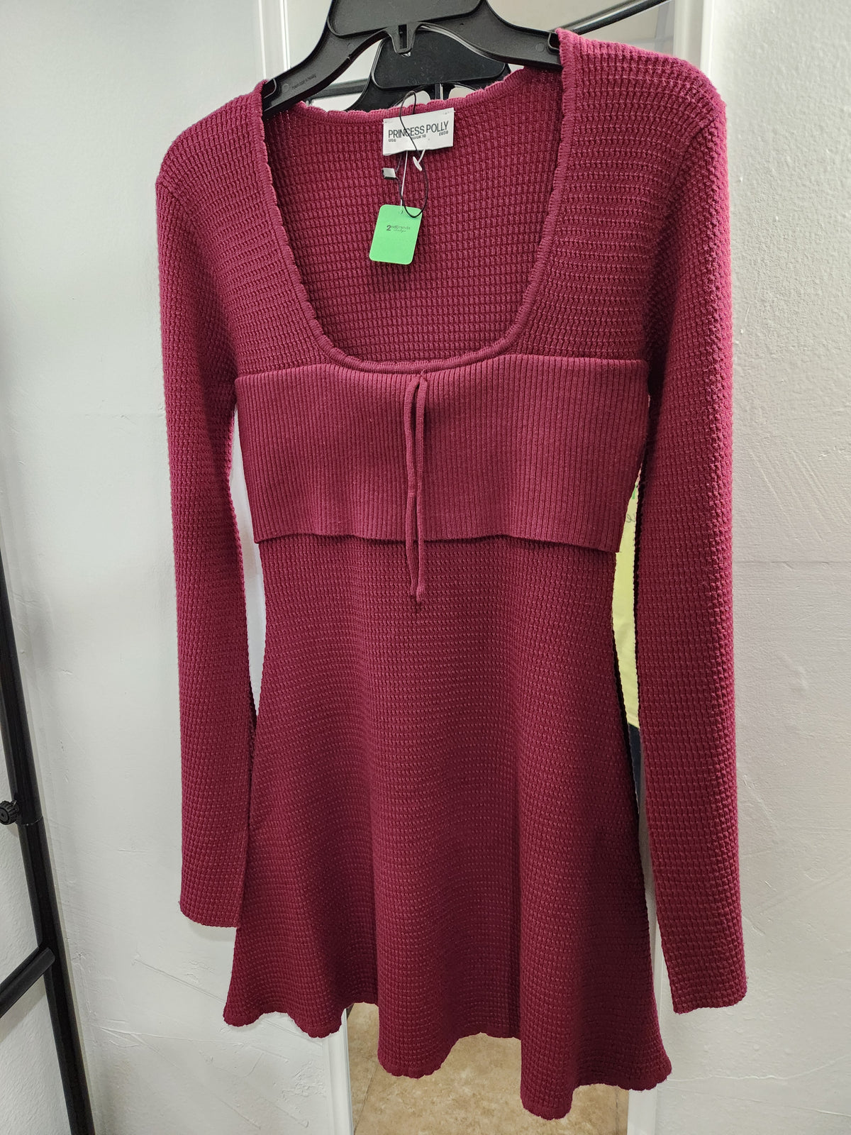 A women's long-sleeve knit dress in a deep berry pink or burgundy, hanging on a black hanger against a white wall. The dress features a scoop neckline, a textured waffle knit pattern, a square waistband with a drawstring detail, and a flared skirt.
