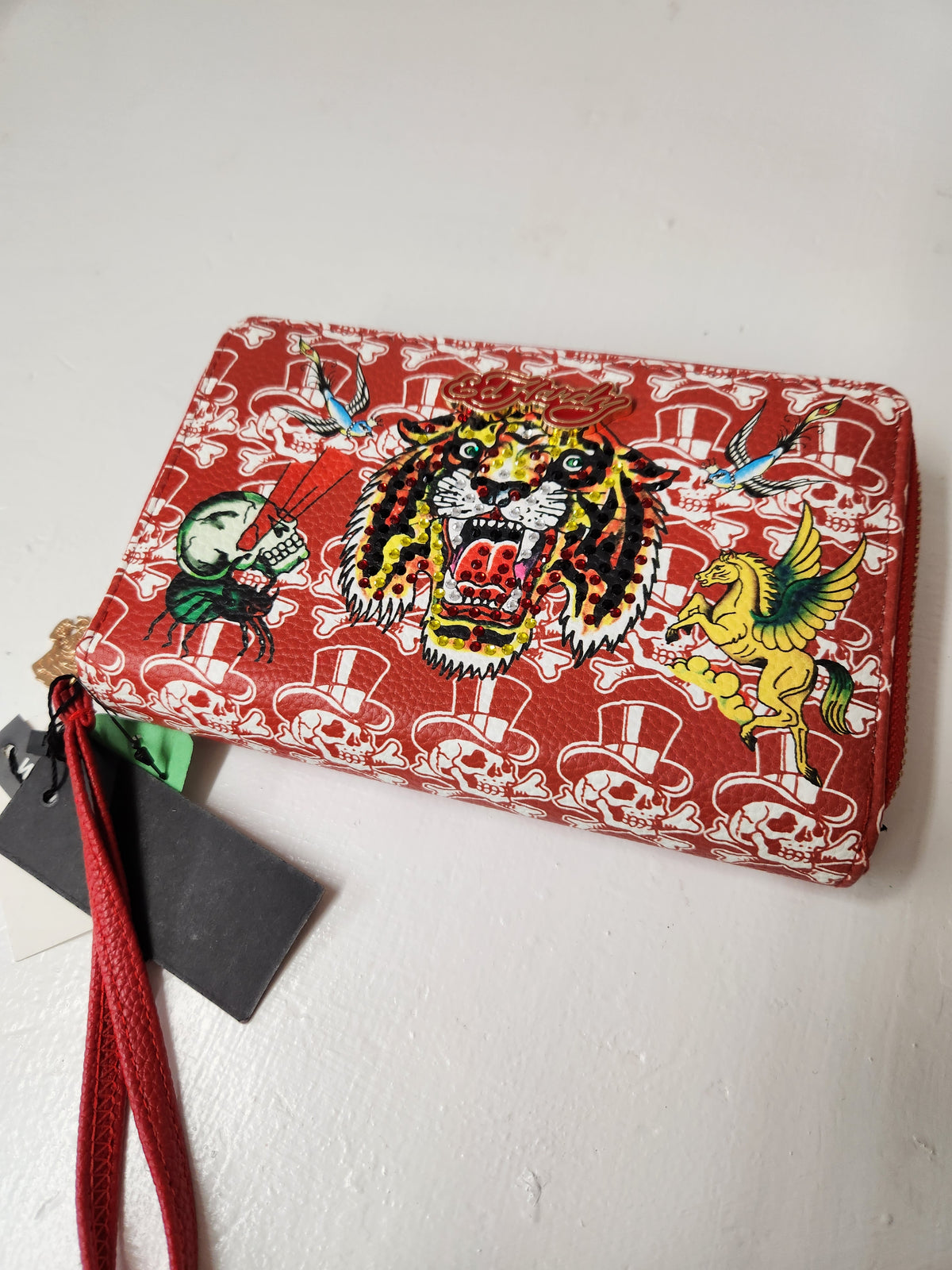  Ed Hardy wristlet wallet features a full zip-around closure, card slot organizers, a zippered coin pocket, and a detachable wristlet strap. Prominent rhinestone-embellished roaring tiger head centered among traditional tattoo flash graphics, including a winged Pegasus and skulls.