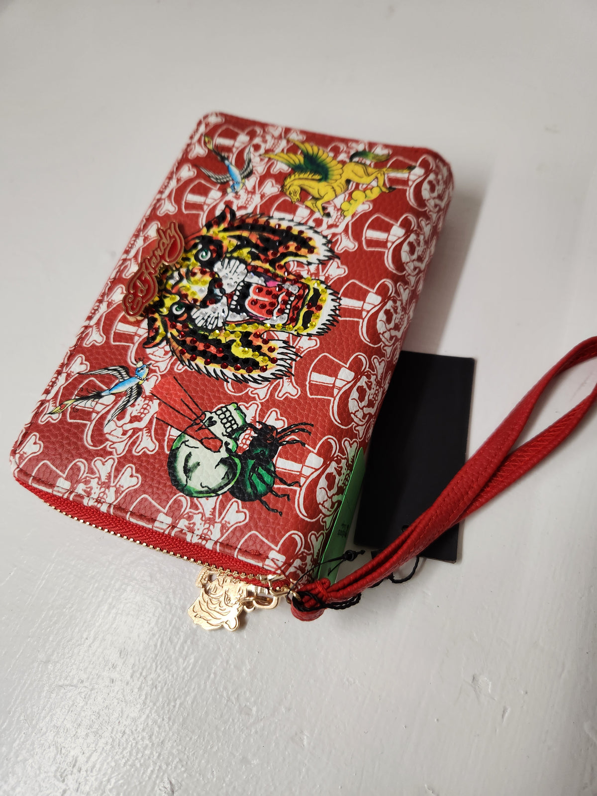  Ed Hardy wristlet wallet features a full zip-around closure, card slot organizers, a zippered coin pocket, and a detachable wristlet strap. Prominent rhinestone-embellished roaring tiger head centered among traditional tattoo flash graphics, including a winged Pegasus and skulls.