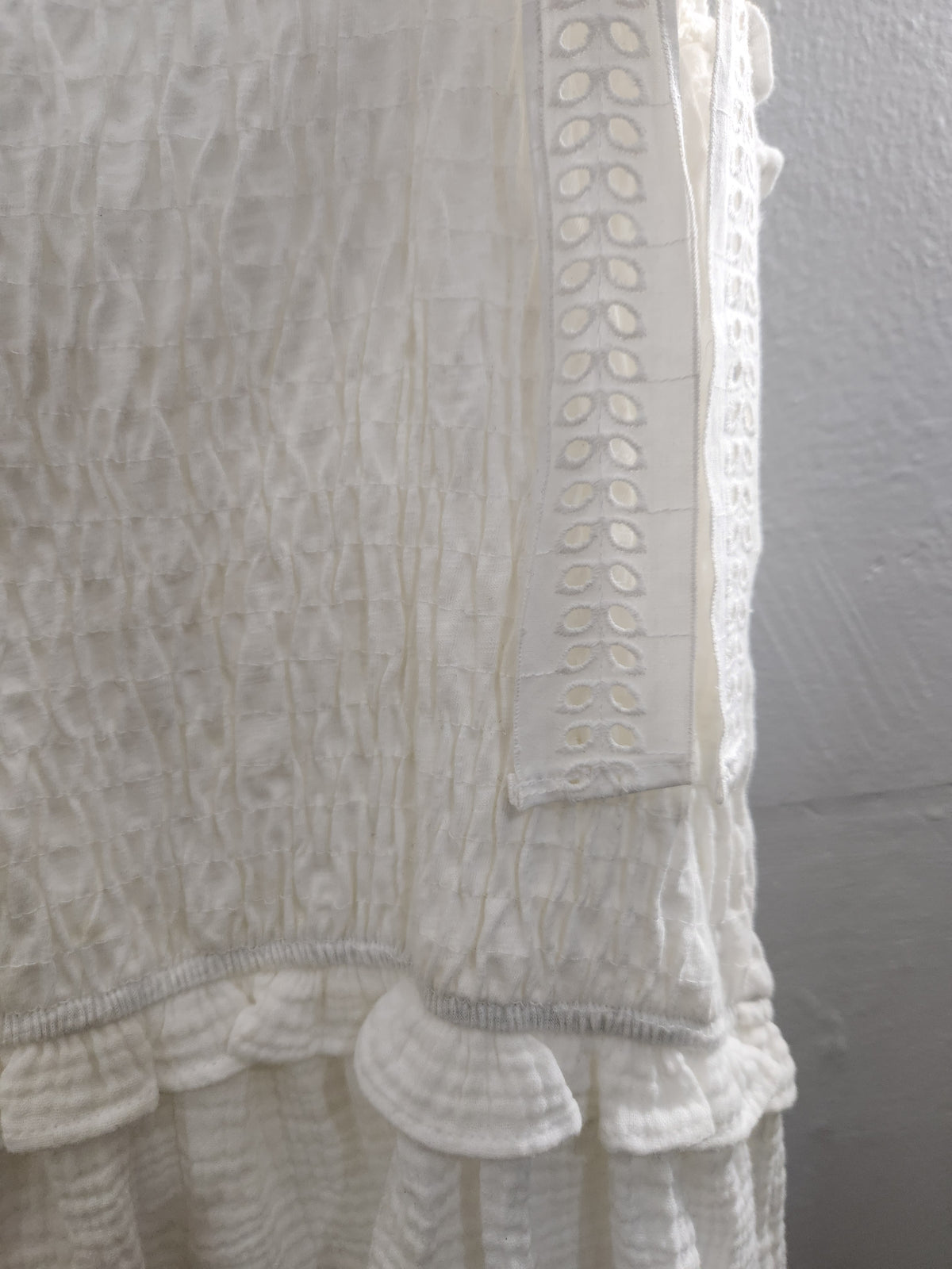 Close-up of a white textured fabric garment with a smocked pattern, eyelet lace trim, and ruffled hem against a light gray wall.