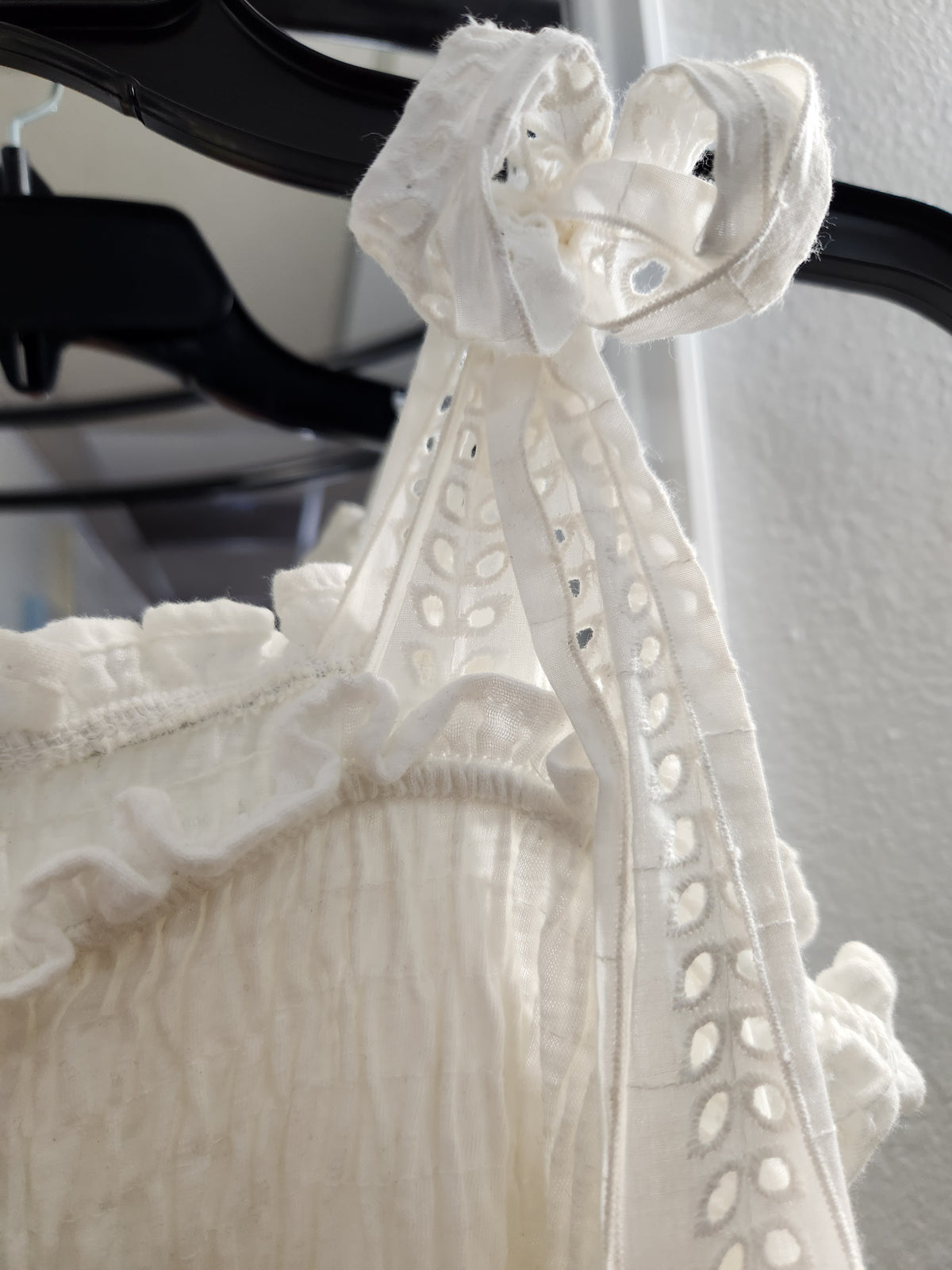 Close-up of a white embroidered lace top hanging on a black hanger against a light wall.