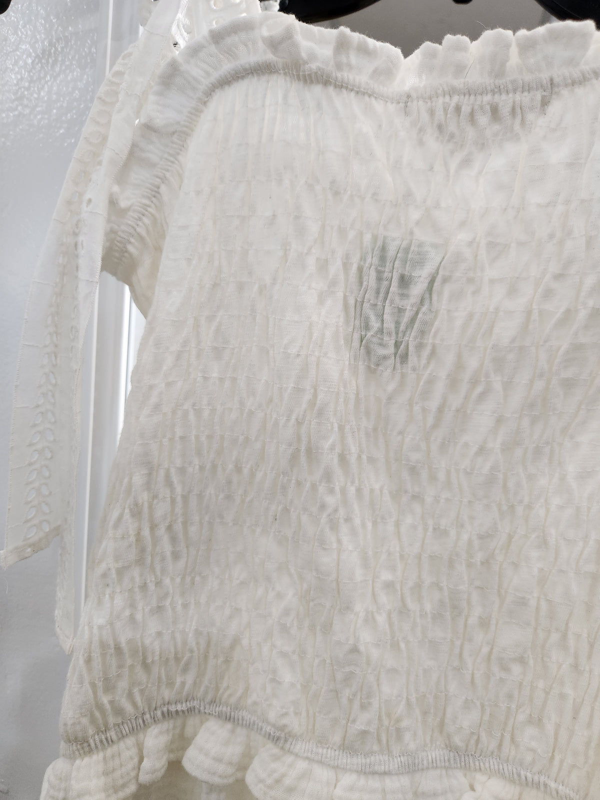 A close-up of a white textured smocked blouse with ruffled trim, hanging on a black hanger against a light background.