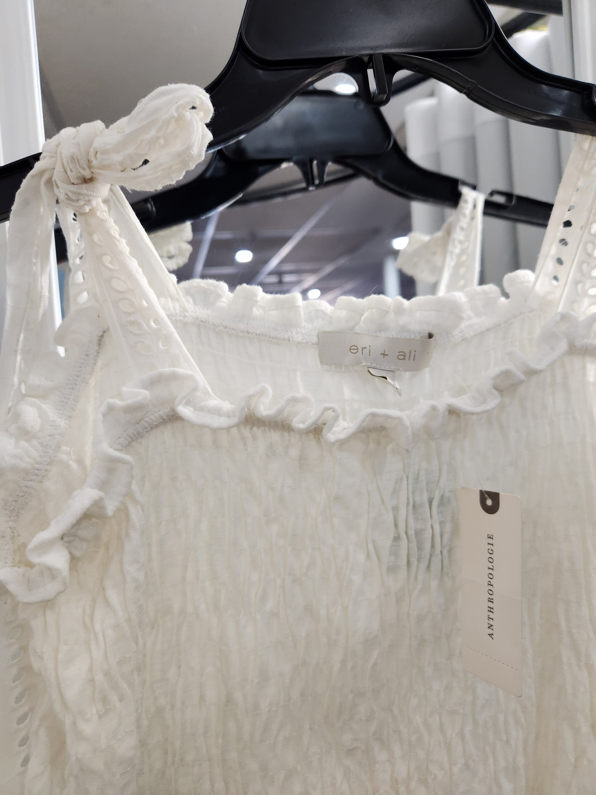 White textured sleeveless top with ruffled trim and tie straps, hanging on a black hanger with a visible brand tag.