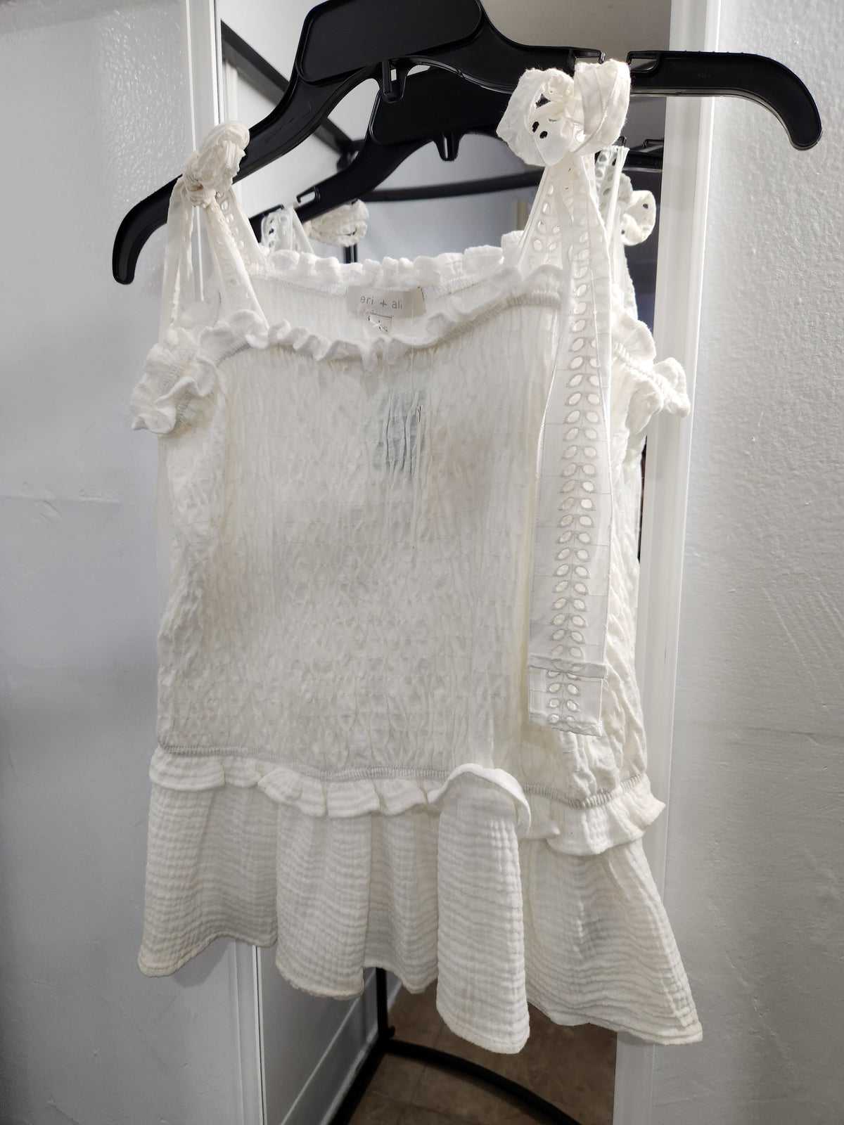 An off-white textured sleeveless top with ruffles and lace-up shoulder ties, hanging on a black hanger against a white door.