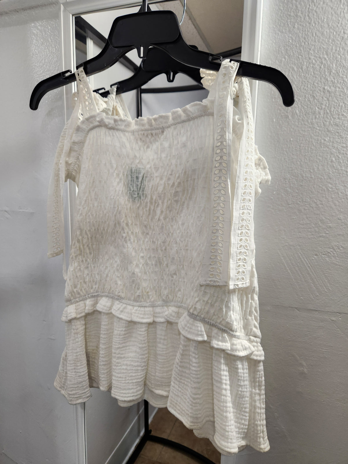 An off-white textured sleeveless top with ruffles and lace trim, hanging on a black hanger against a white wall.
