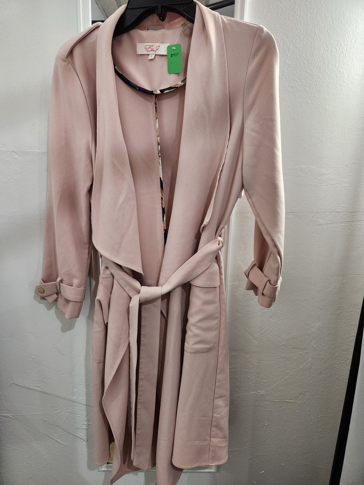 A light pink longline wrap coat with a tie belt, hanging on a black hanger against a light gray wall. A patterned blouse and a green price tag are visible underneath.