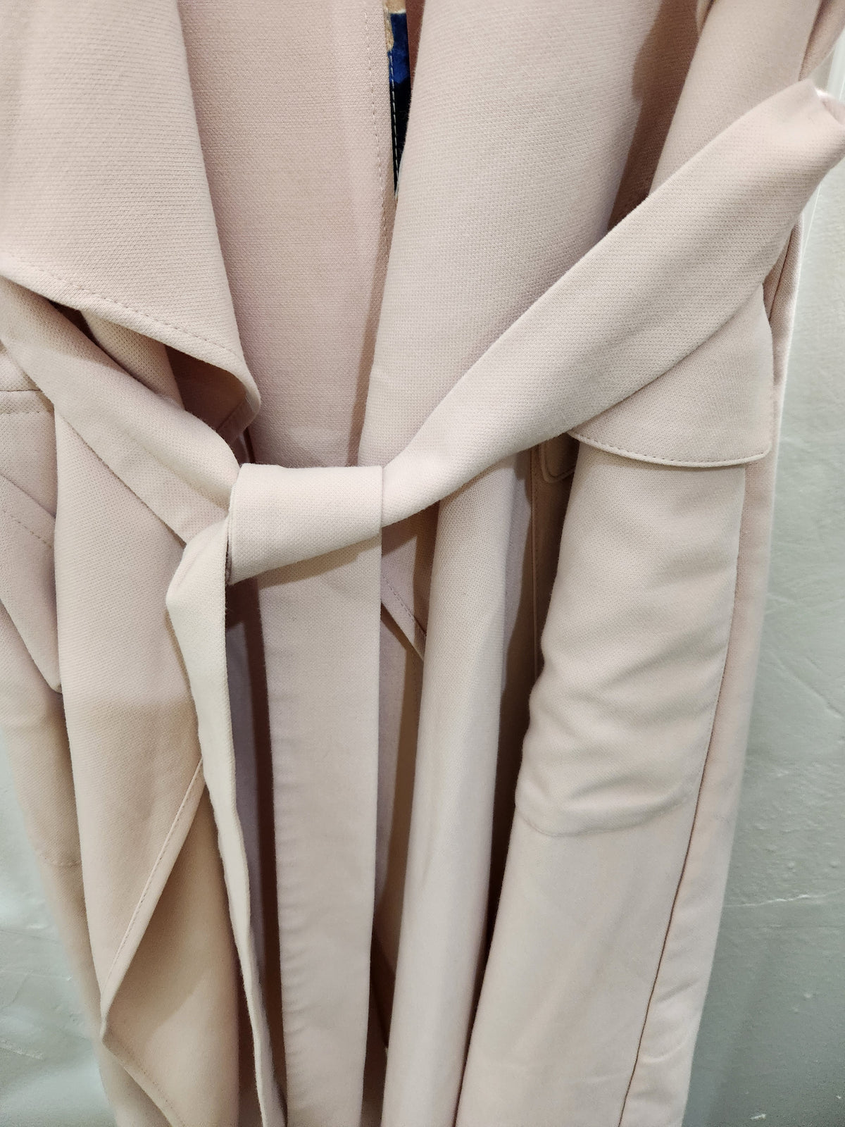 A close-up of a light beige or cream-colored wrap coat or jacket, showing the fabric texture and a tie belt detail.