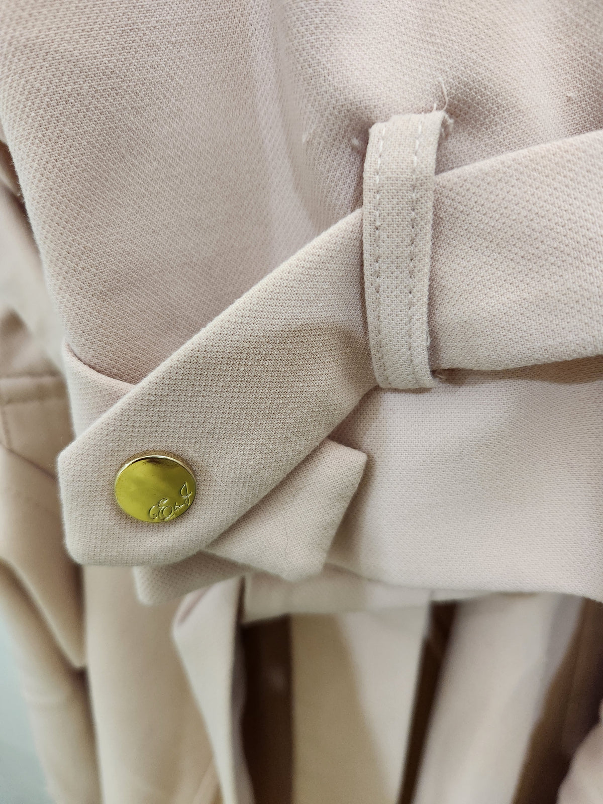 Close-up of a beige trench coat with a matching waist belt and a gold-tone button closure.