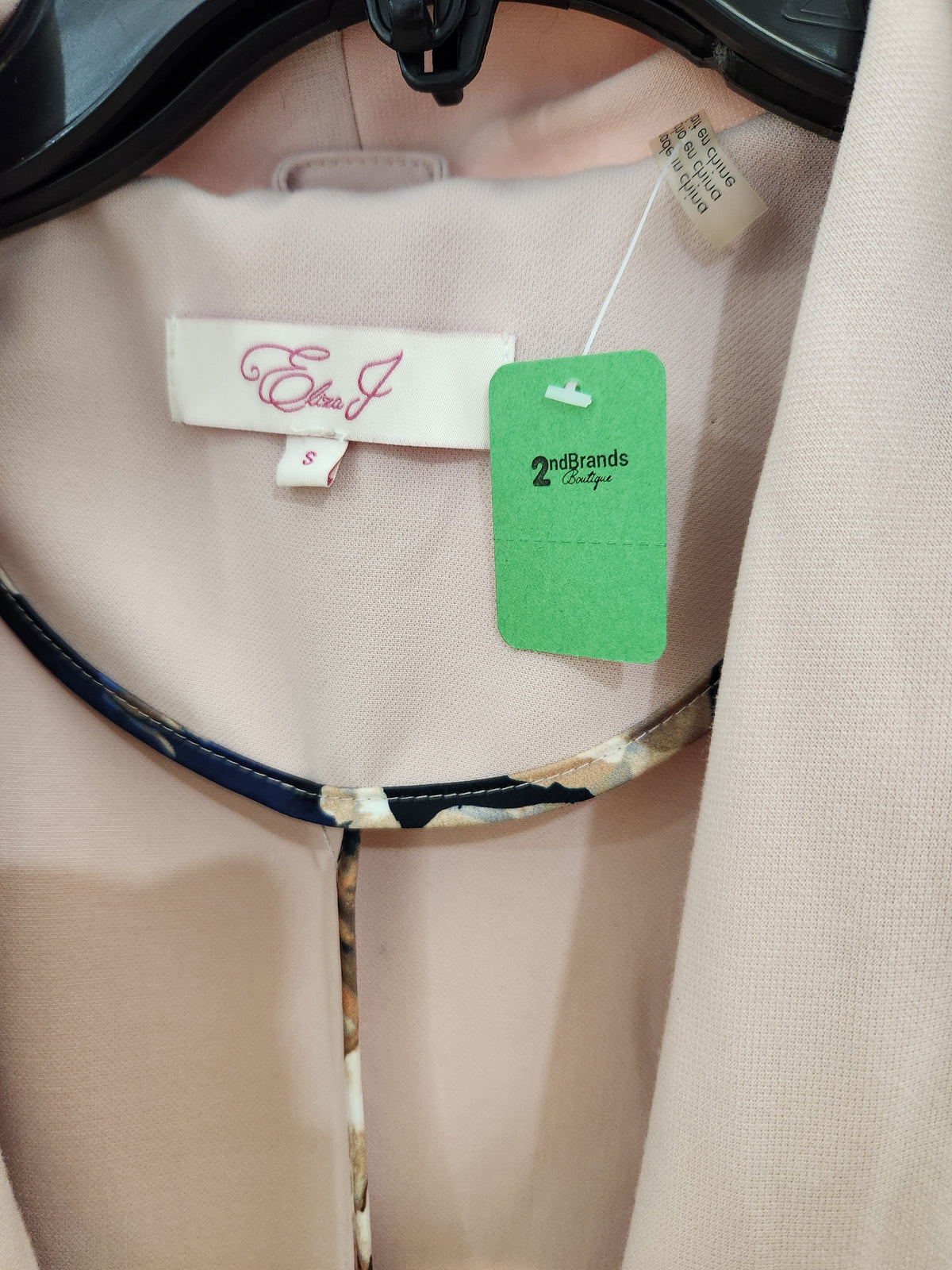 A close-up of a light pink blouse on a hanger, showing the interior neck label with 'Elena J' in pink script and a green '2brands Boutique' tag.