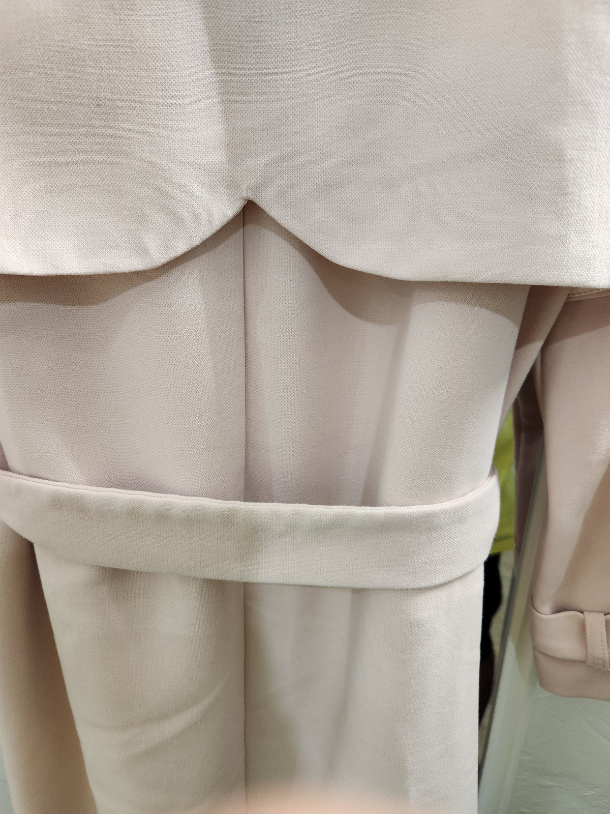 Close-up of a light beige or cream-colored garment, showing the back of a cropped jacket or coat with a wide waist belt and matching trousers.