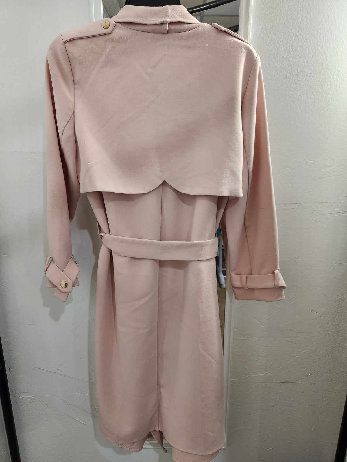 A light pink trench coat dress displayed on a hanger against a light gray wall. The long-sleeved dress features a structured collar, shoulder epaulets, a matching waist belt, and buttoned cuffs.
