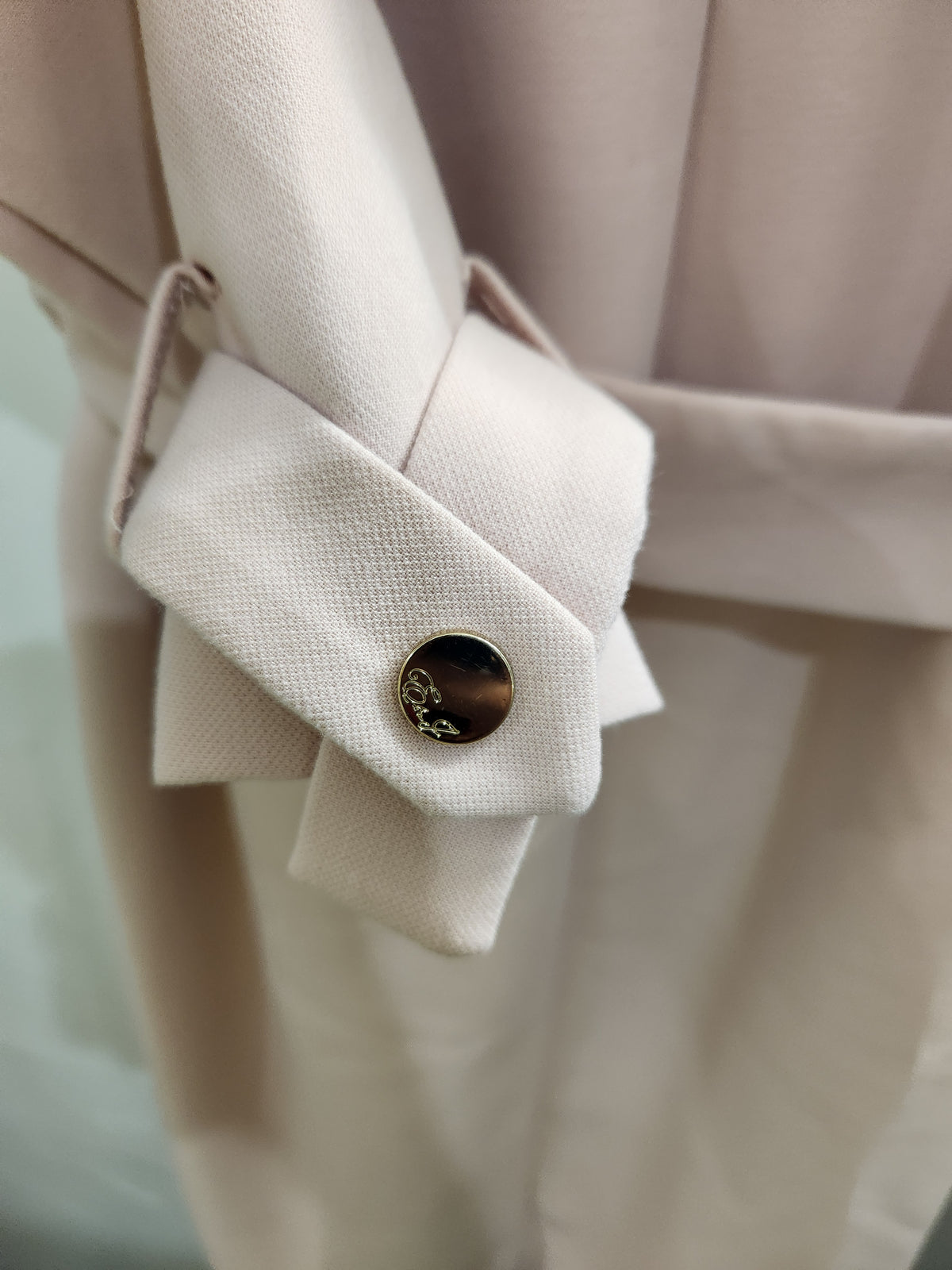 Close-up of a light beige or cream-colored fabric garment with a structured cuff and a round dark metallic button featuring a small gold-toned floral detail.
