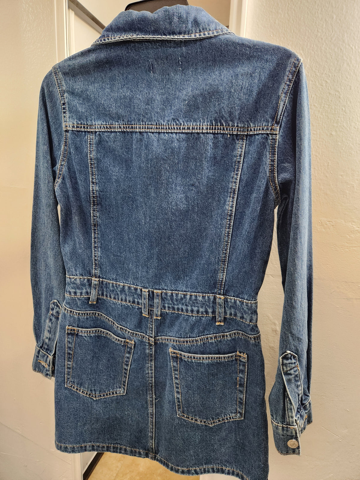 A blue denim jacket with a belted waist and front patch pockets, shown from the back on a mannequin against a beige wall.