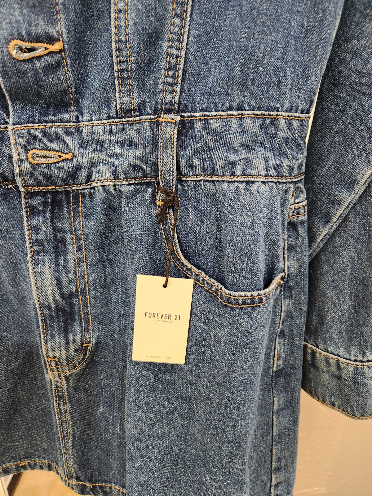 Close-up of a blue denim garment with visible front pocket, contrast tan stitching, and a hanging Forever 21 tag.