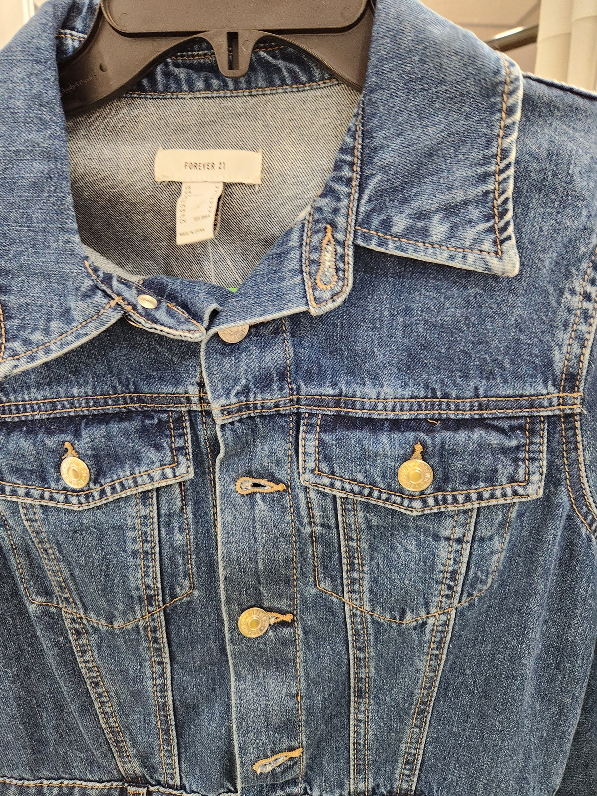 A close-up of a blue denim jacket from Forever 21 hanging on a black hanger. The jacket features a classic collar, button-up front, and two buttoned chest pockets with gold-tone buttons.