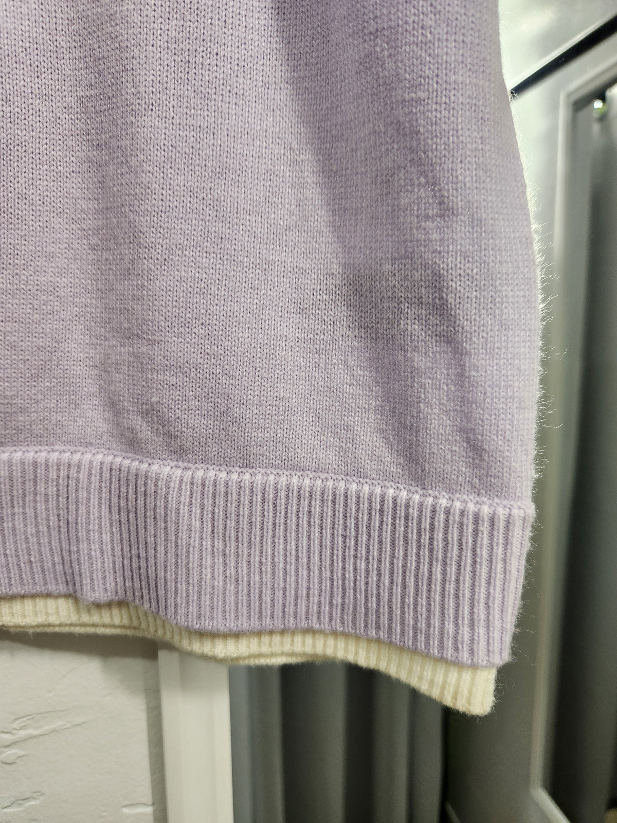 Close-up of a light lavender knit garment with a ribbed hem, showing a cream-colored inner lining or second ribbed trim along the edge.