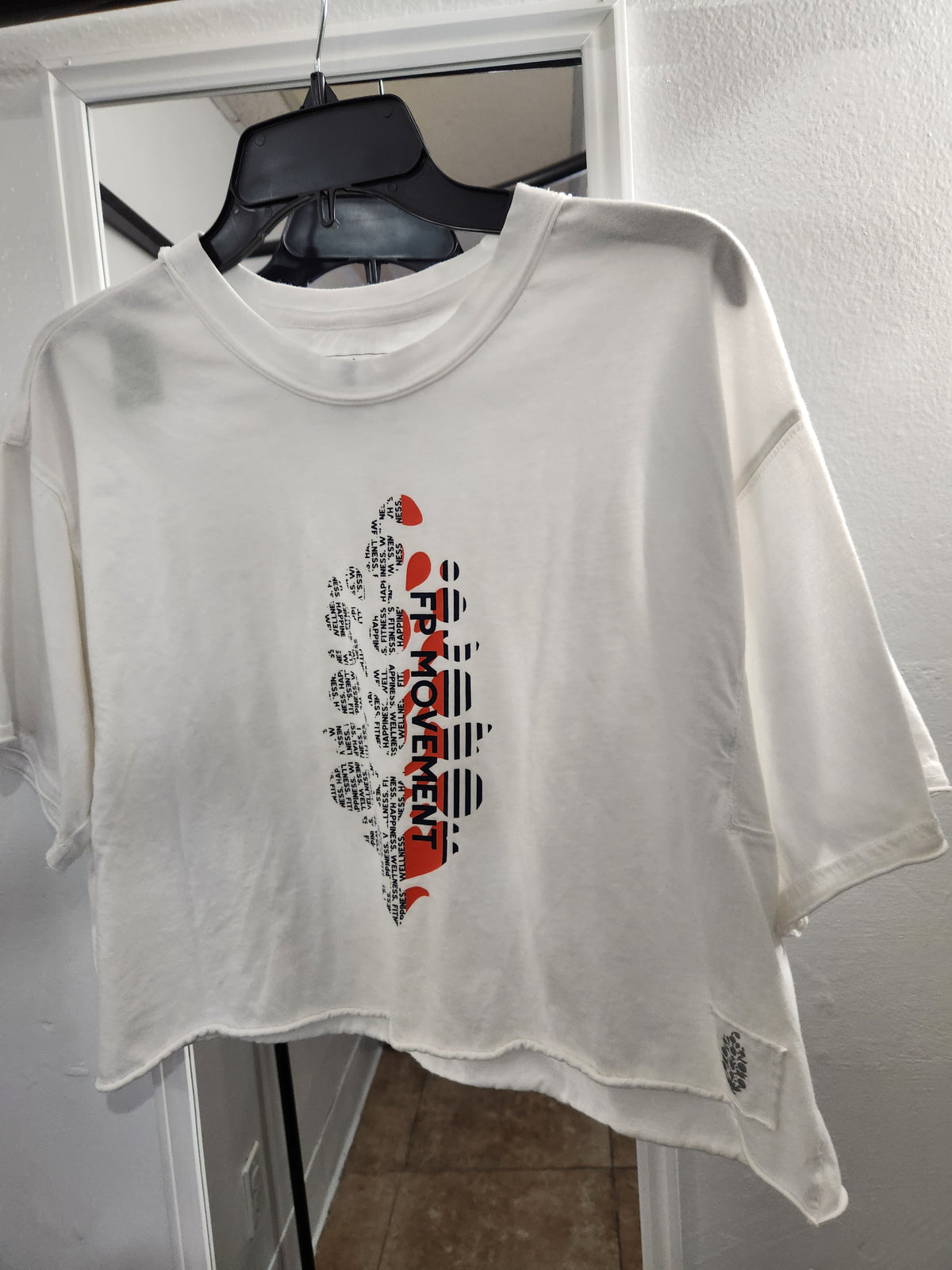 A white cropped t-shirt with a distressed, frayed hem and sleeve edges, featuring a graphic print of the state of Florida filled with text and red accents. The shirt is hanging on a black hanger in front of a white door.