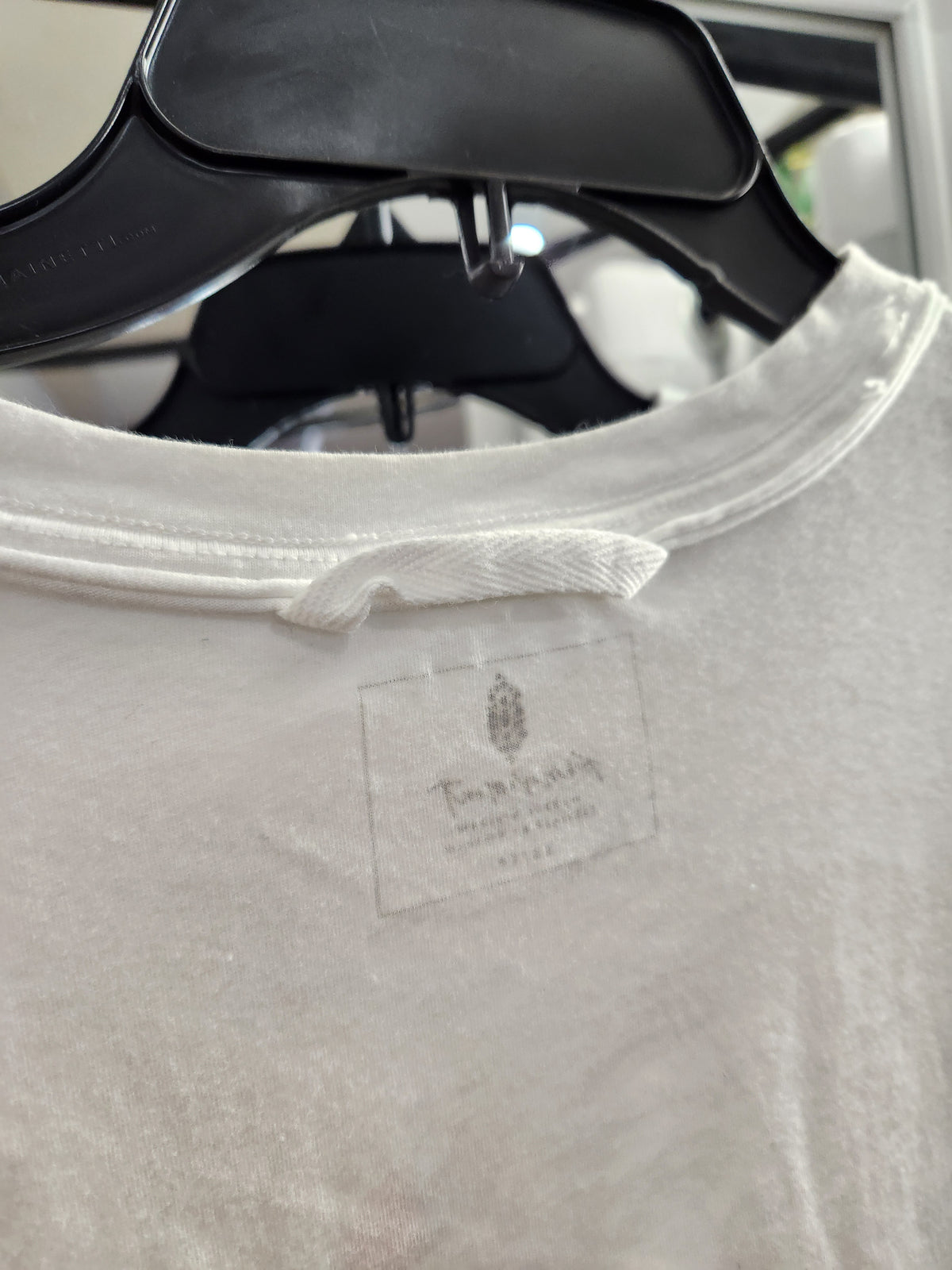 Close-up of a white crewneck t-shirt hanging on a black plastic hanger, showing a faintly visible brand label on the inside neck.