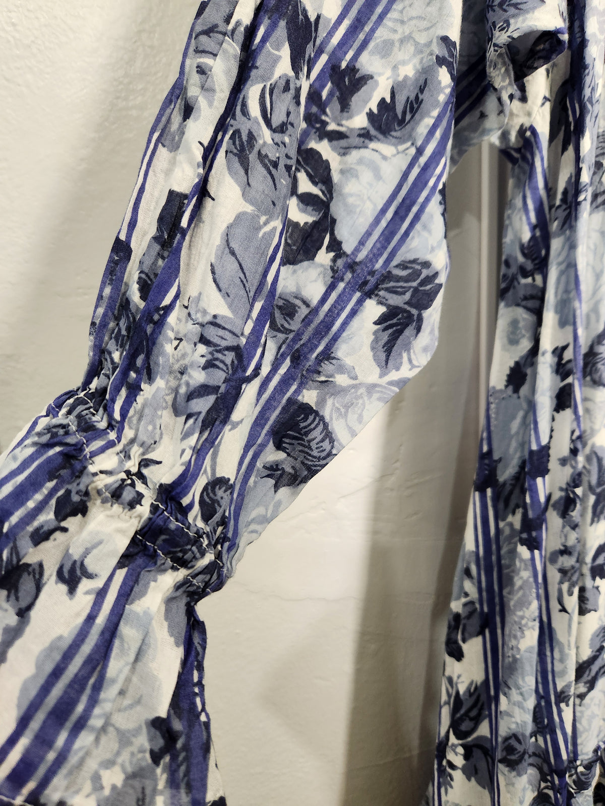 A close-up of a blue and white floral print fabric with vertical stripes and gathered elastic details, shown hanging against a white wall.