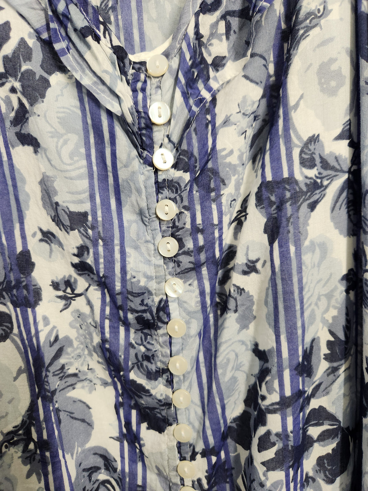 A close-up of a button-front blouse with a blue and gray floral print over a white base, featuring vertical blue stripes and white buttons.