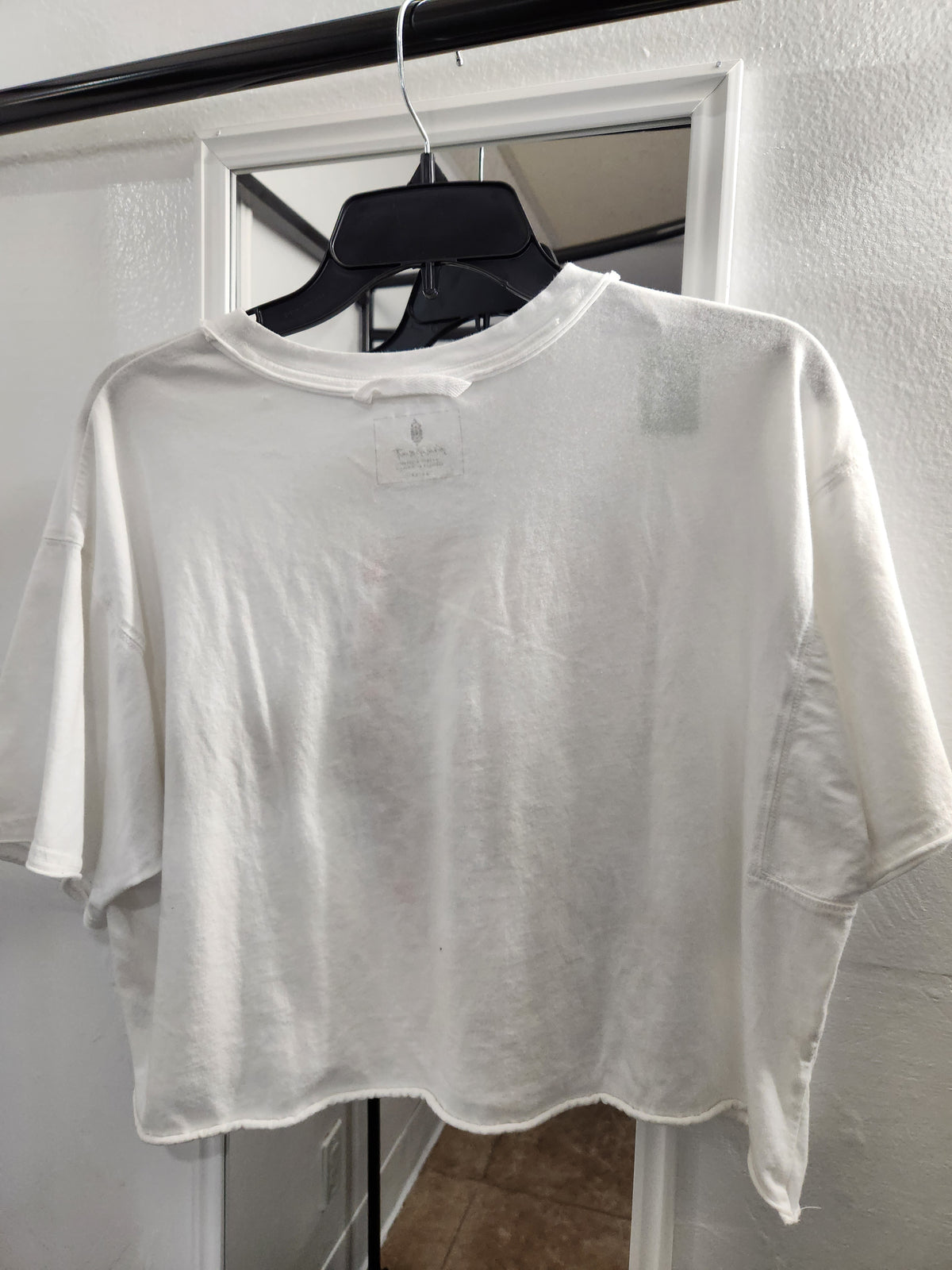A white oversized short-sleeve t-shirt hanging on a black hanger from a clothing rack, positioned in front of a white door and textured wall.