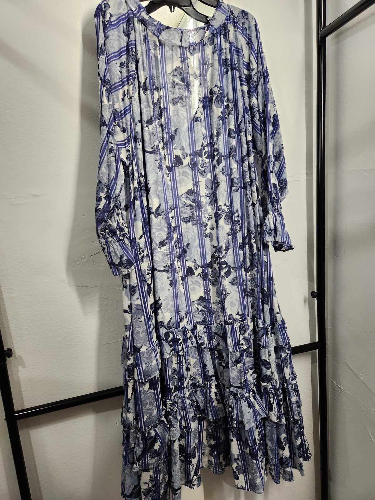 A long-sleeve midi dress with a blue and white toile-style floral print, hanging on a black hanger against a white wall.