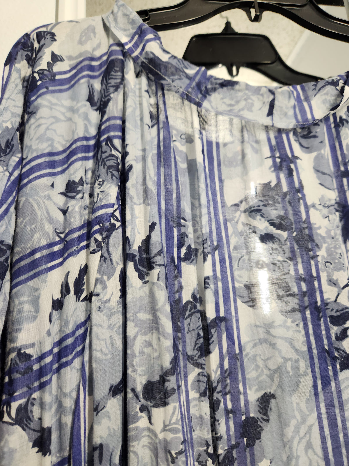 A close-up of a lightweight blouse hanging on a black hanger, featuring a blue and gray floral print over a white base with vertical stripes.