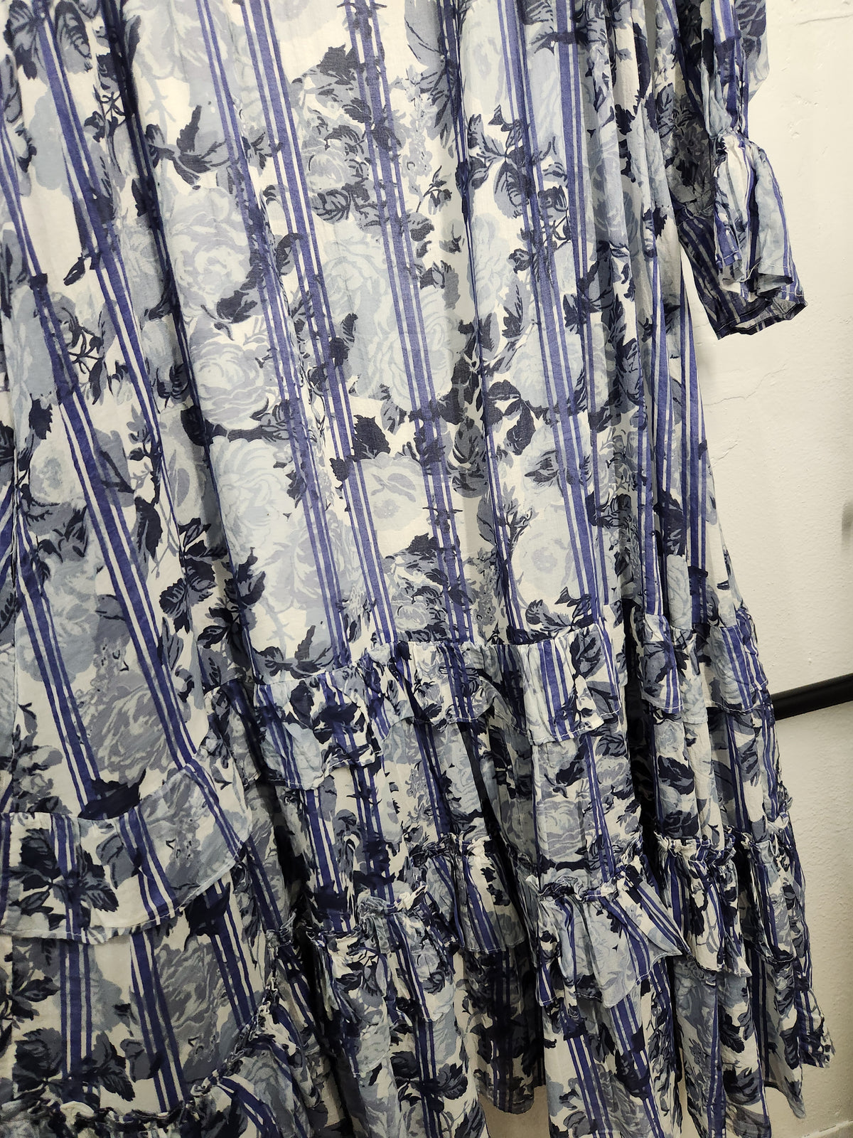 A tiered maxi dress with a blue and white floral print, featuring vertical stripes and ruffled details, shown hanging on a rack.
