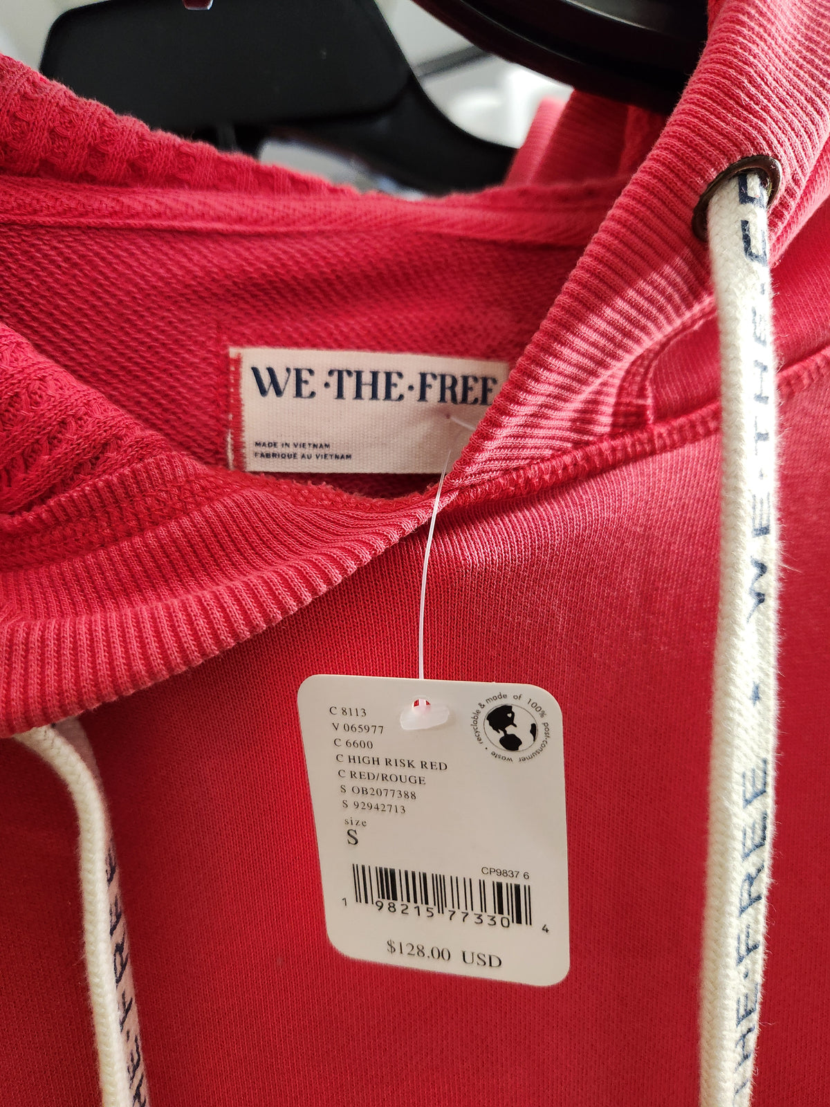 A close-up of a red hoodie with white drawstrings and a visible neck label reading WE'THEFREE, with a product tag hanging from the collar.