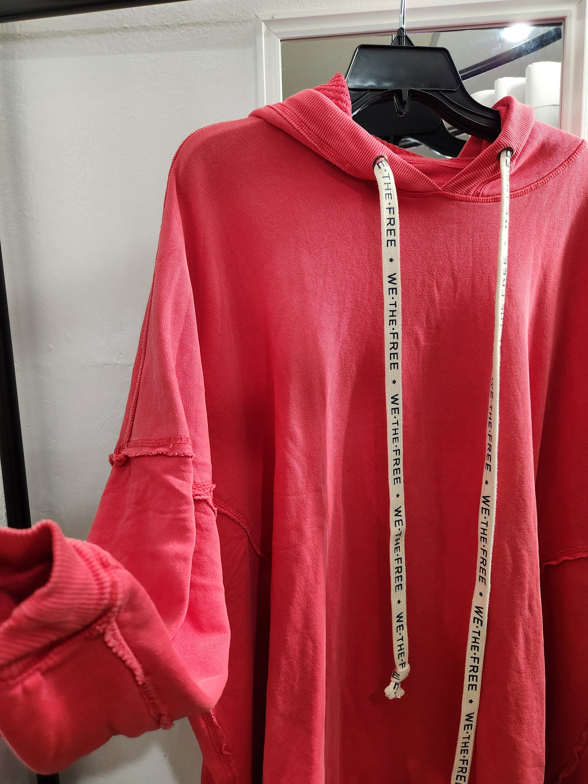 A coral pink hooded sweatshirt hanging on a black hanger against a white wall. The hoodie has distressed details on the sleeves, a white drawstring with black text reading 'WE THE FREE', and a matching white drawstring hanging down the front.