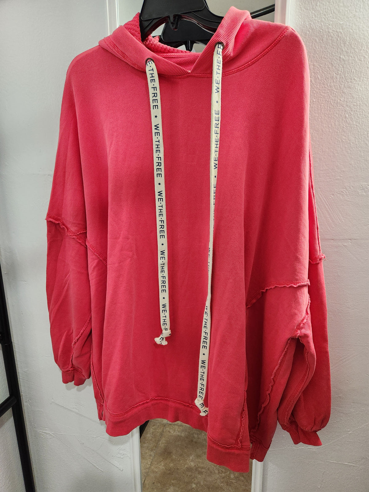 A faded pink distressed hoodie hanging on a black hanger against a white wall. The hoodie features white drawstrings printed with the text 'WE THE FREE WE THE FREE'.