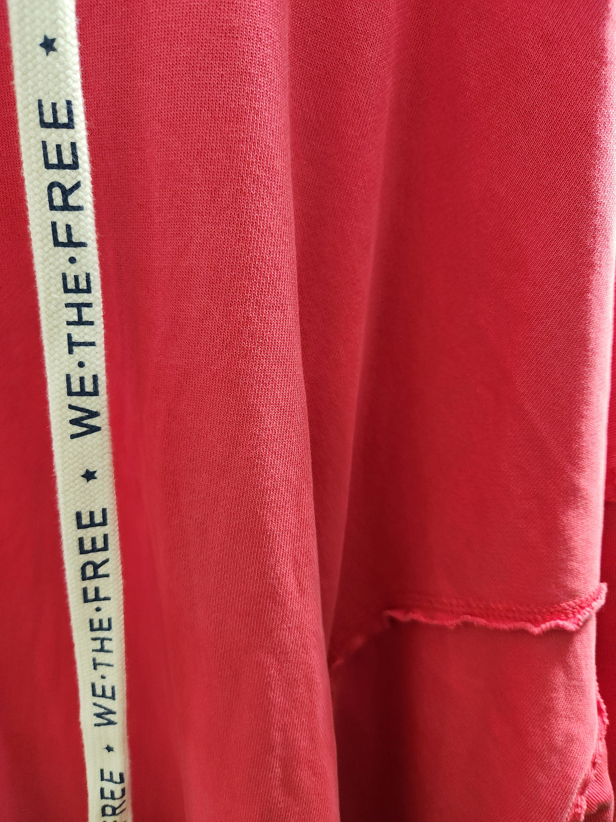 Close-up of a red fabric garment with a vertical white stripe printed with the words 'WE·THE·FREE' in black, plus a small torn hem detail.